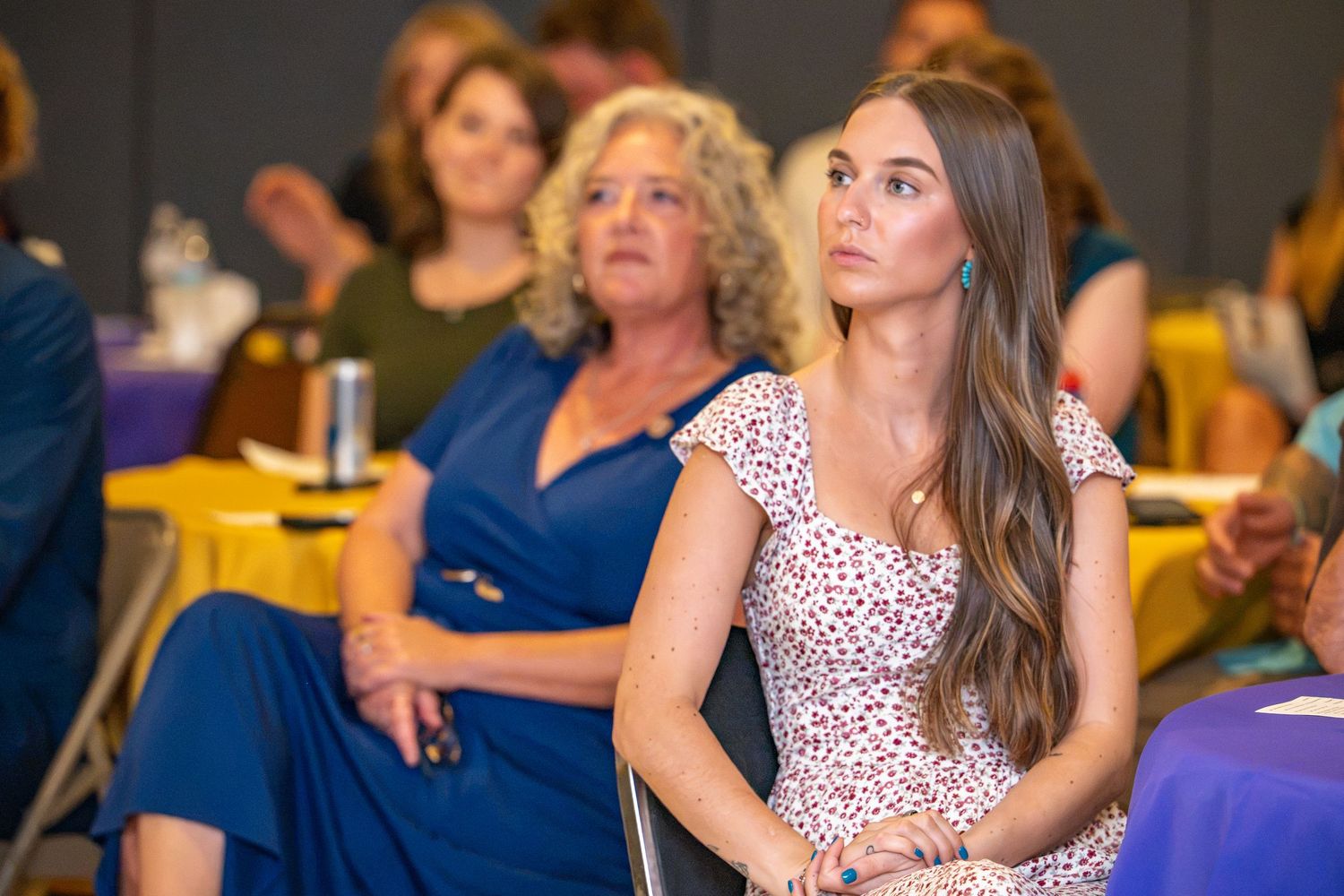 Two women seated at an event, one in a blue dress and one in a floral dress, attentively watching a presentation.