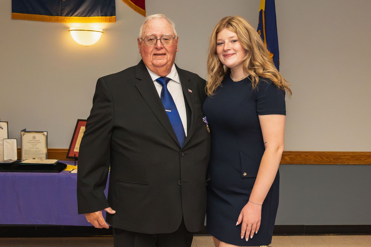 Elderly man in suit and young blonde woman in navy dress posing together at formal event.