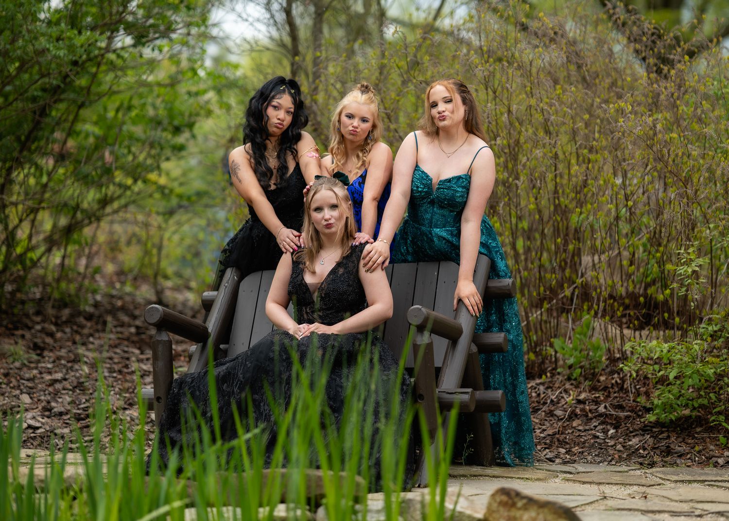 Four young women in formal gowns pose on a garden bench surrounded by lush greenery for prom photos.