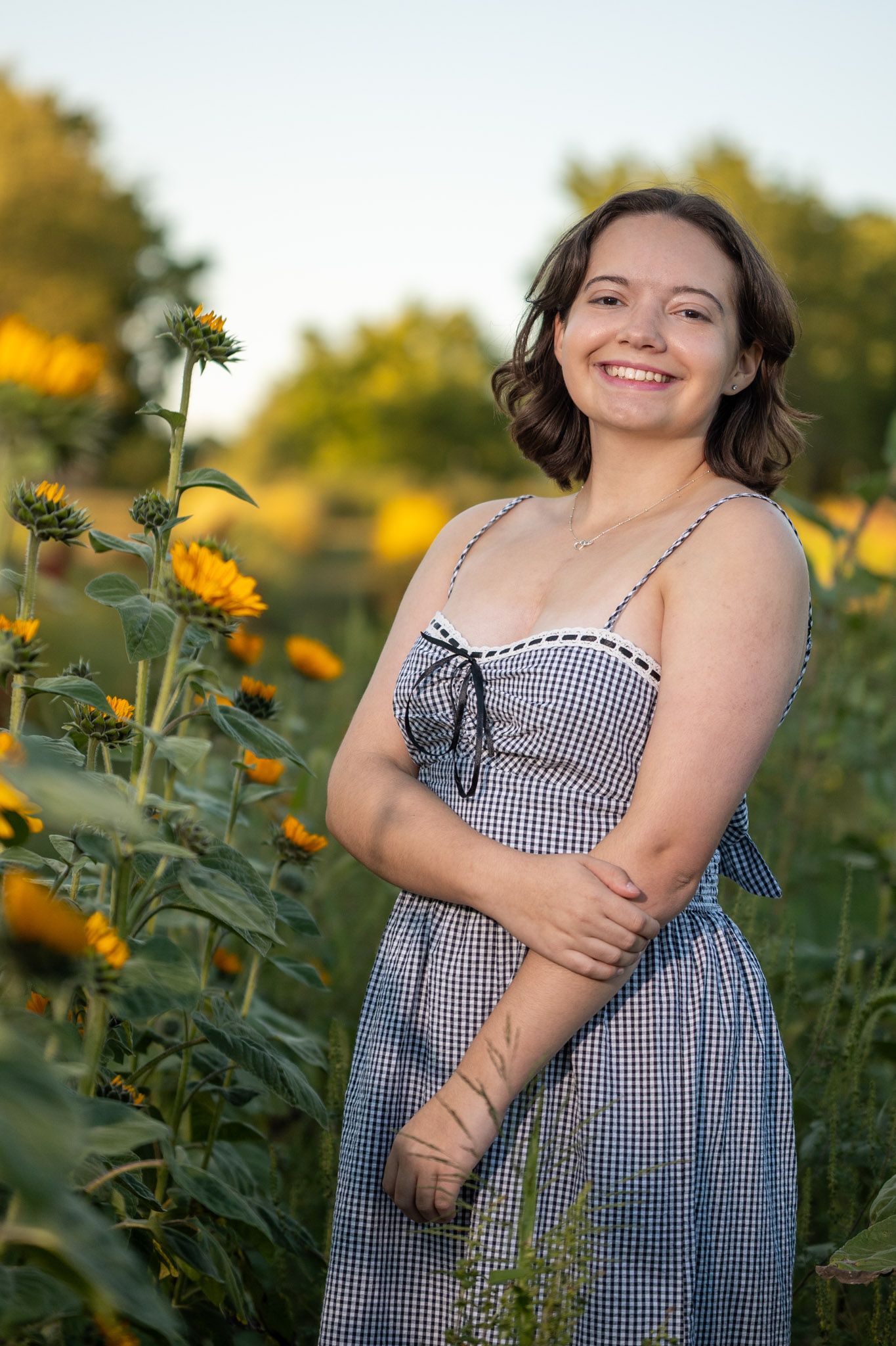 A gingham dress-wearing figure stands joyfully in a field of bright yellow sunflowers on a sunny summer day.