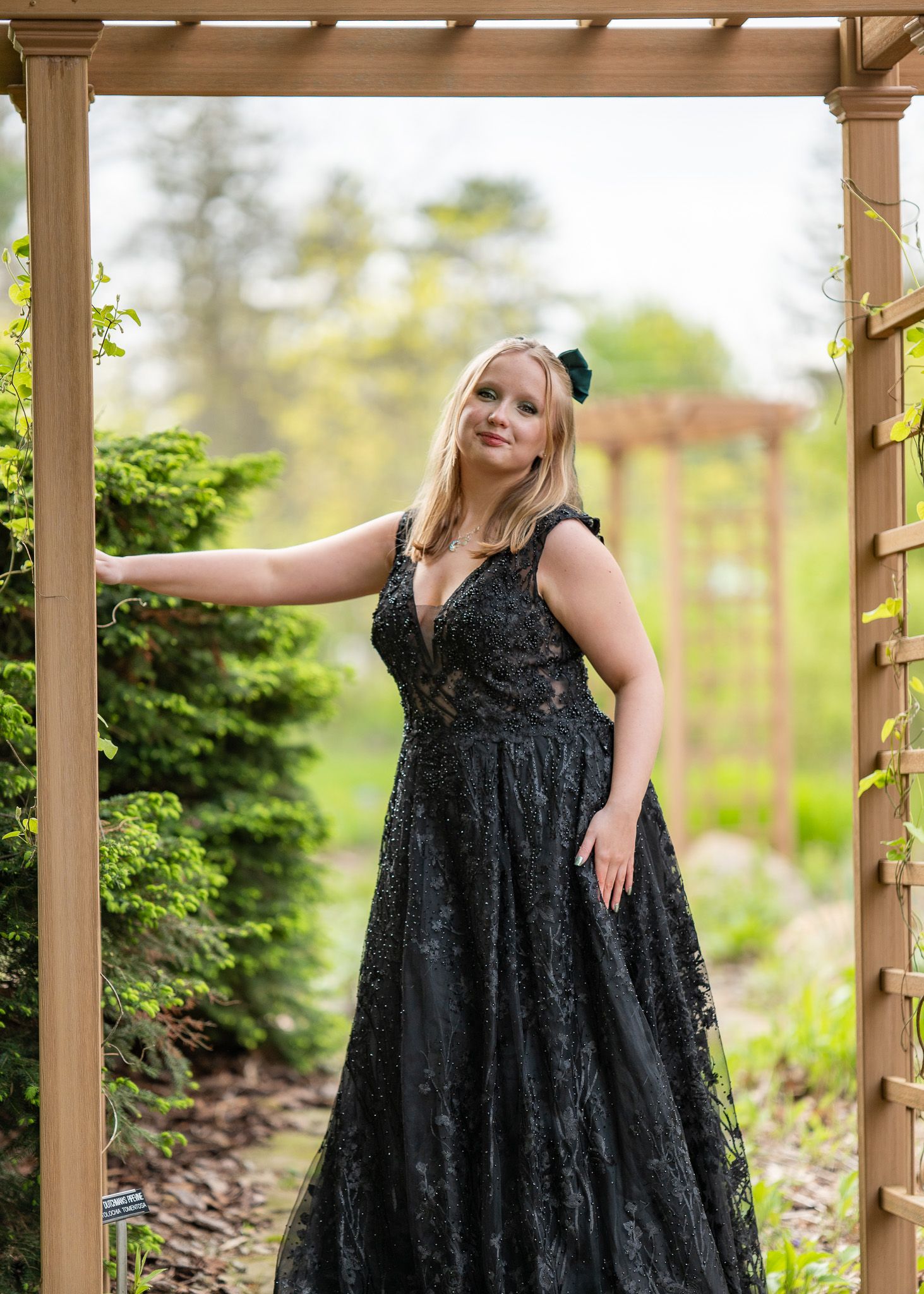 Blonde woman in black lace ball gown poses under wooden garden arbor surrounded by lush green foliage.