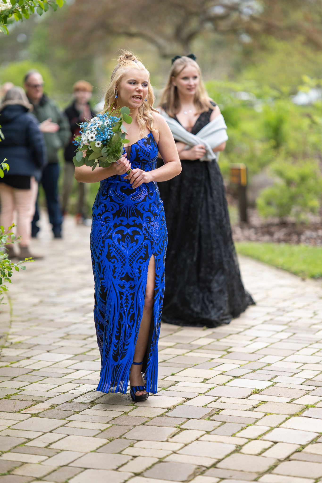 Woman in blue fringe gown holds bouquet on stone path, with guests in formal attire at outdoor wedding.