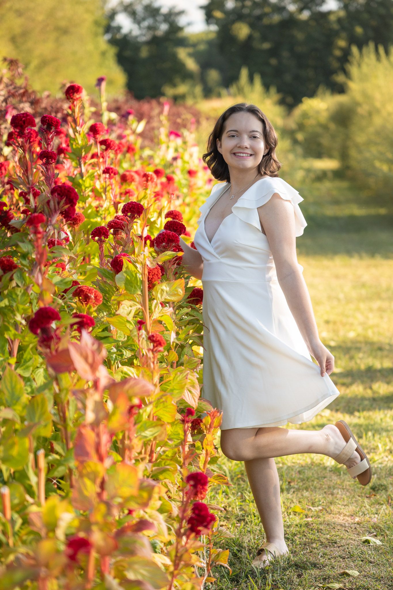 A person in a white dress poses playfully among vibrant red flowers in a sunlit garden.