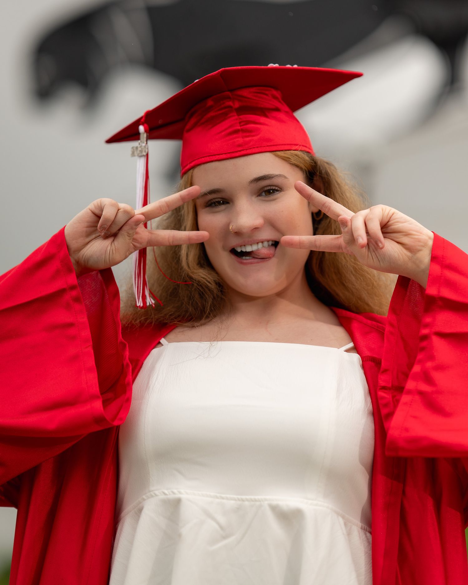 Smiling graduate in red cap and gown making peace signs, wearing white dress underneath at graduation ceremony.