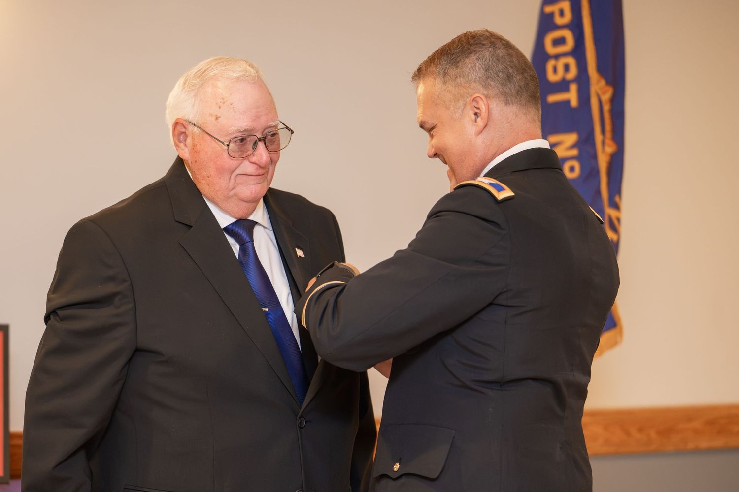 Military officer pins medal on elderly veteran in dark suit at American Legion post ceremony.