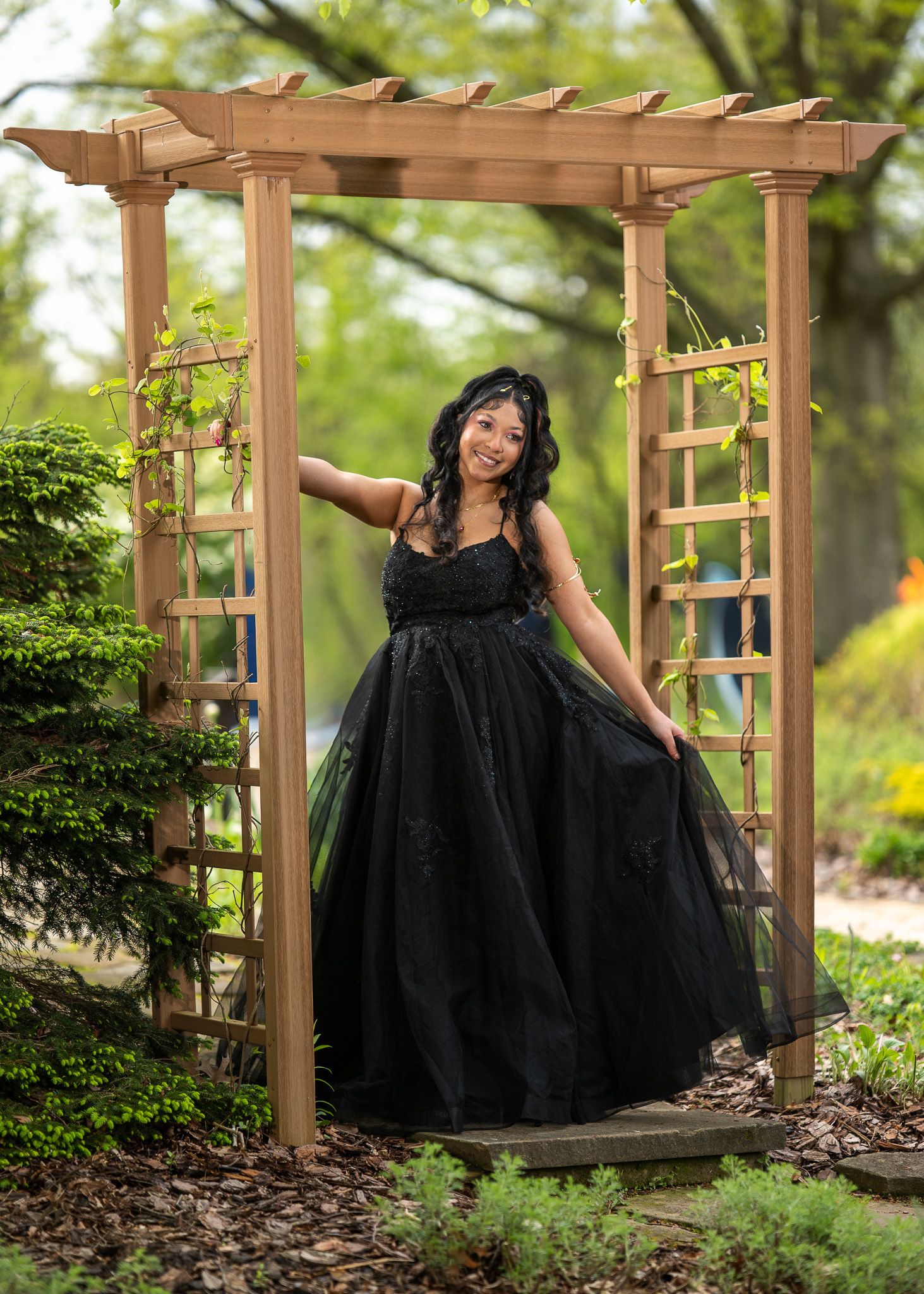 Young woman in elegant black ball gown posing under a wooden garden arbor surrounded by lush greenery.