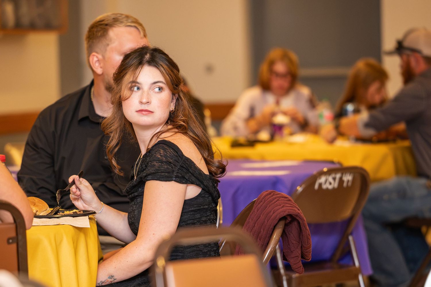 Young brunette woman in black off-shoulder top sitting at table during indoor event.