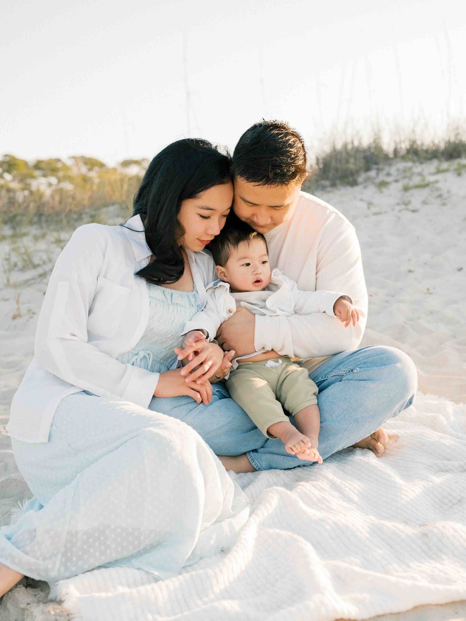 Mom, dad and baby son sitting on a blanket and snuggling during a beach session in Dauphin Island by family photographer, AFV Photography