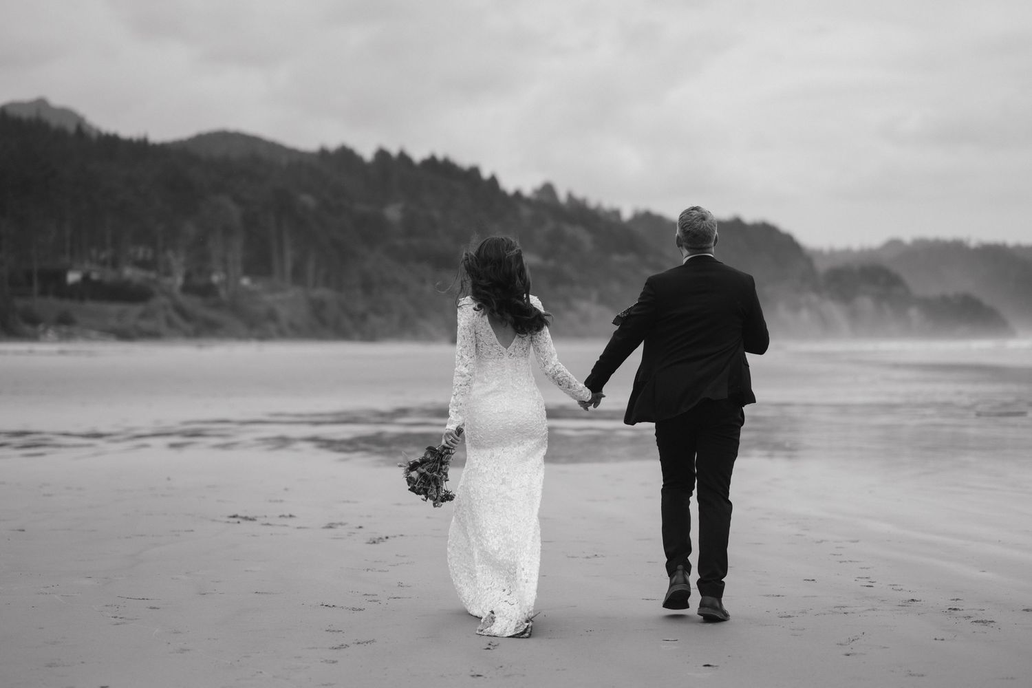 Black and white image of newlyweds walking hand in hand on a beach.