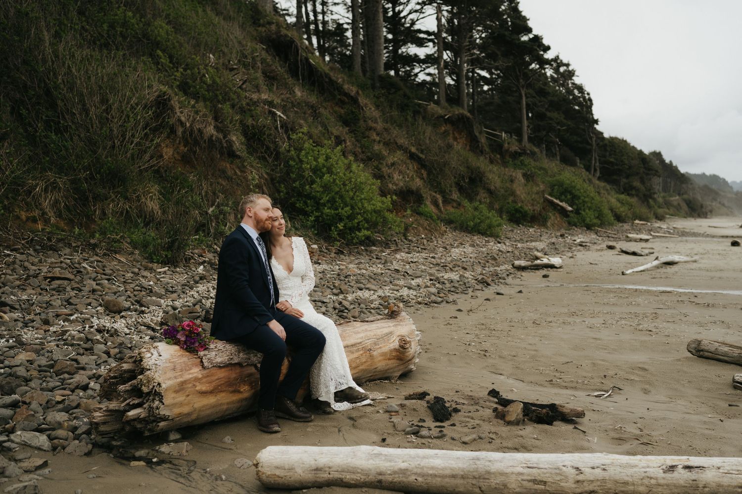 A couple sitting together on driftwood at a moody coastal beach with cliffs in the background.