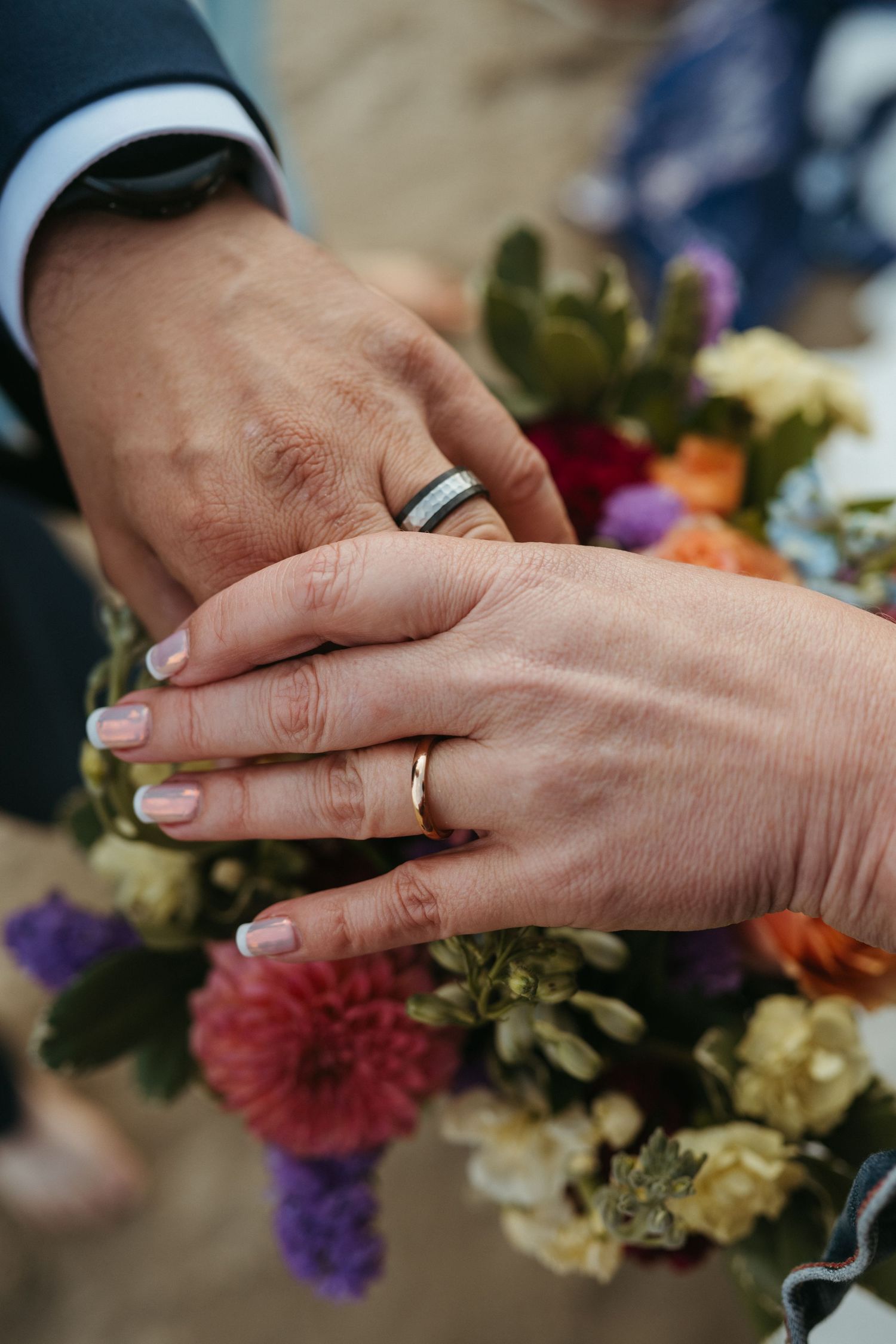 Close-up of wedding rings and hands with colorful floral bouquet in background.