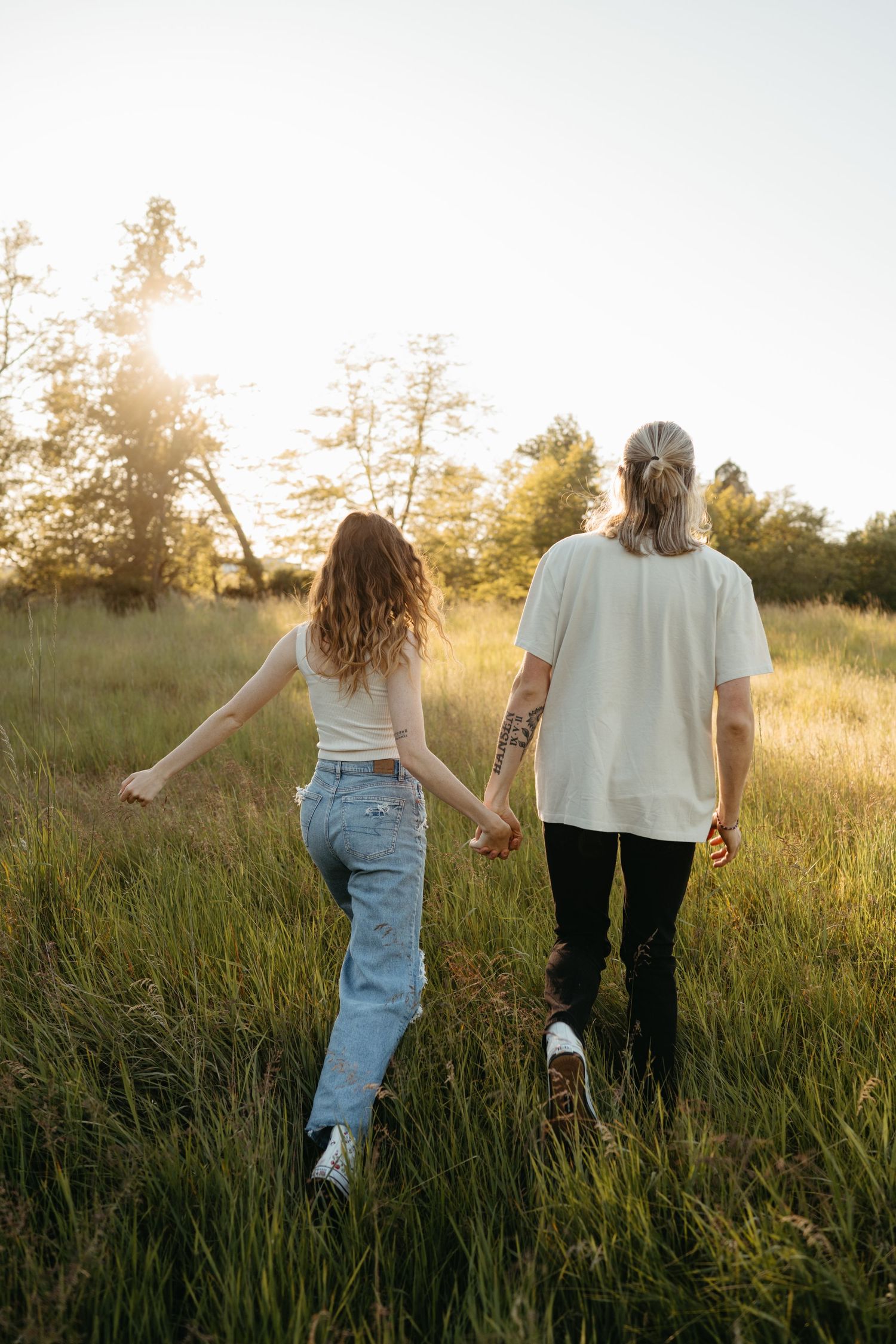 A couple walks through tall grass in a sunlit field during golden hour.