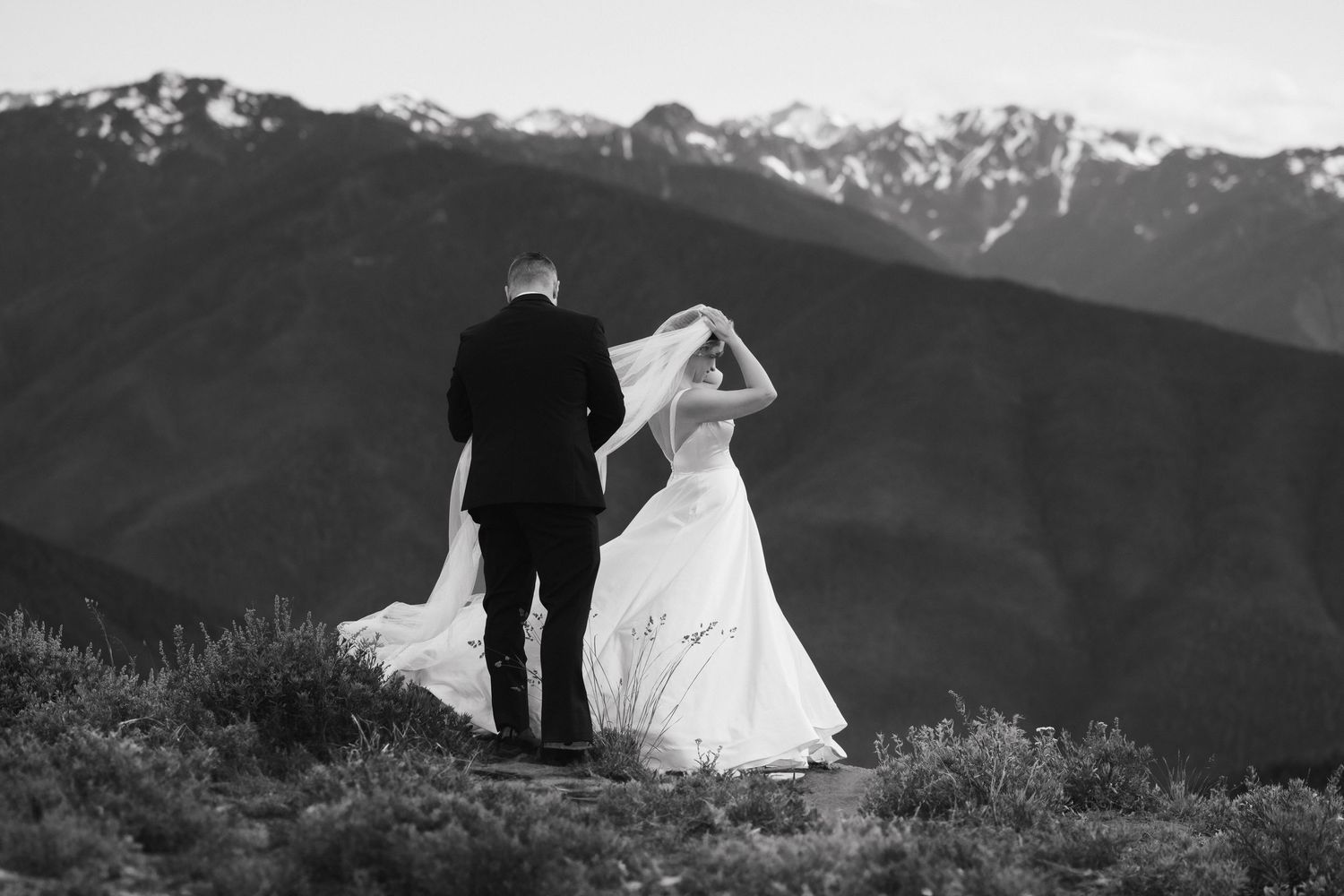Couple stands silhouetted against mountain range as bride's dress flows in wind.