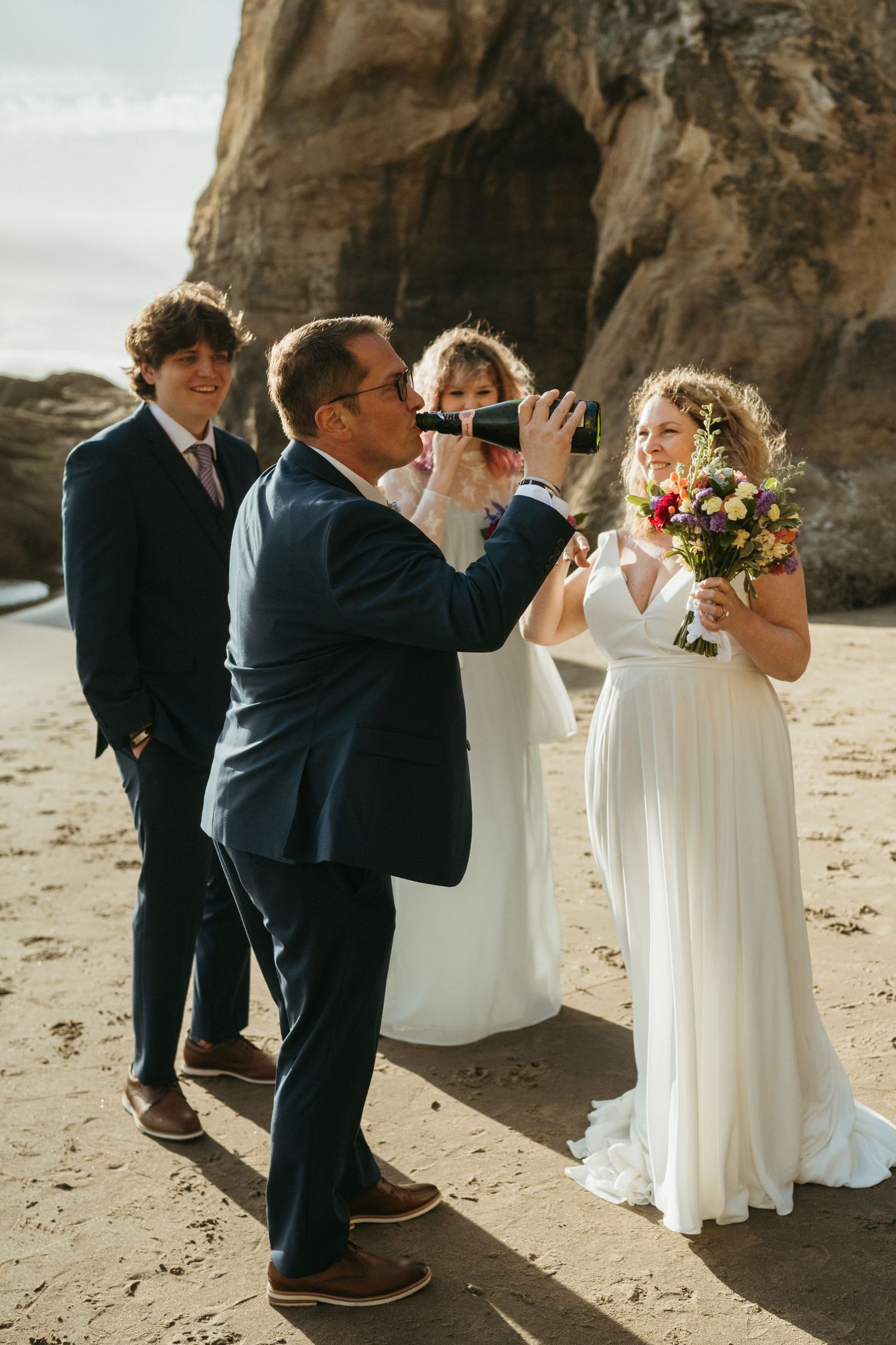 A wedding photographer captures moments during a beach ceremony near rocky cliffs.