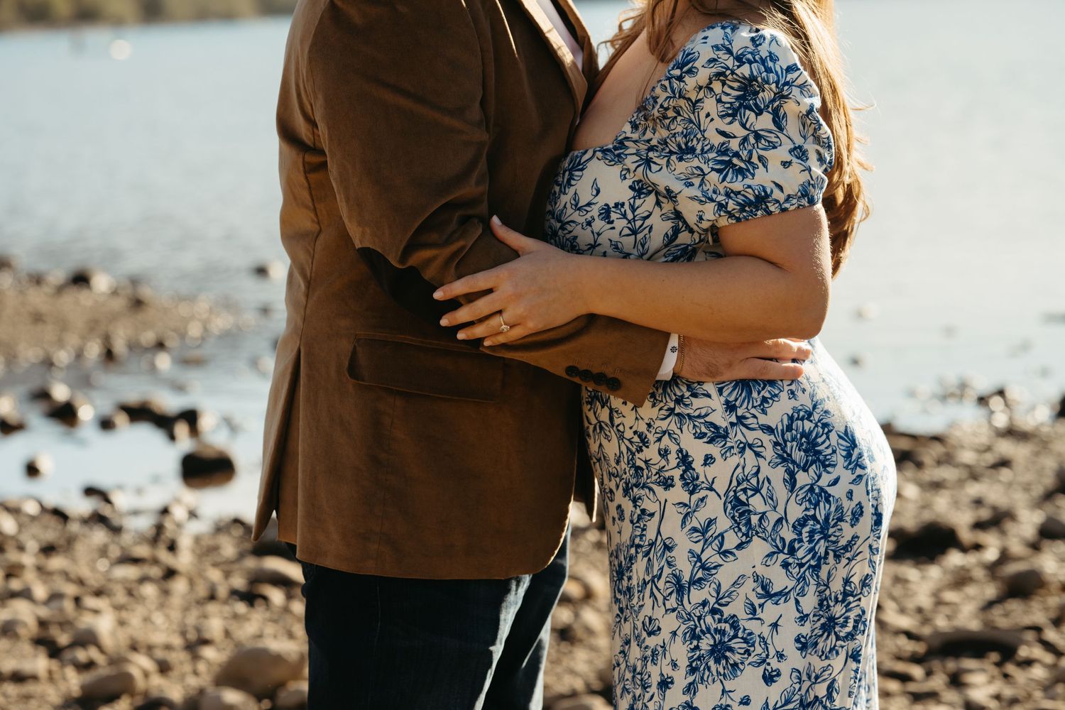 A couple in formal attire embraces on a rocky shoreline with sunlit water in the background.
