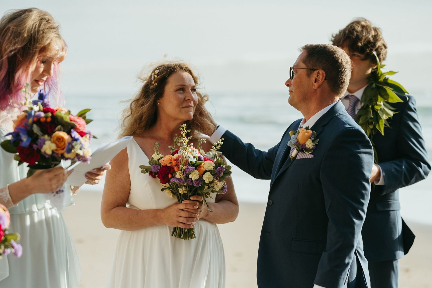 Wedding ceremony participants share joyful moment with colorful bouquets on sunny beach.