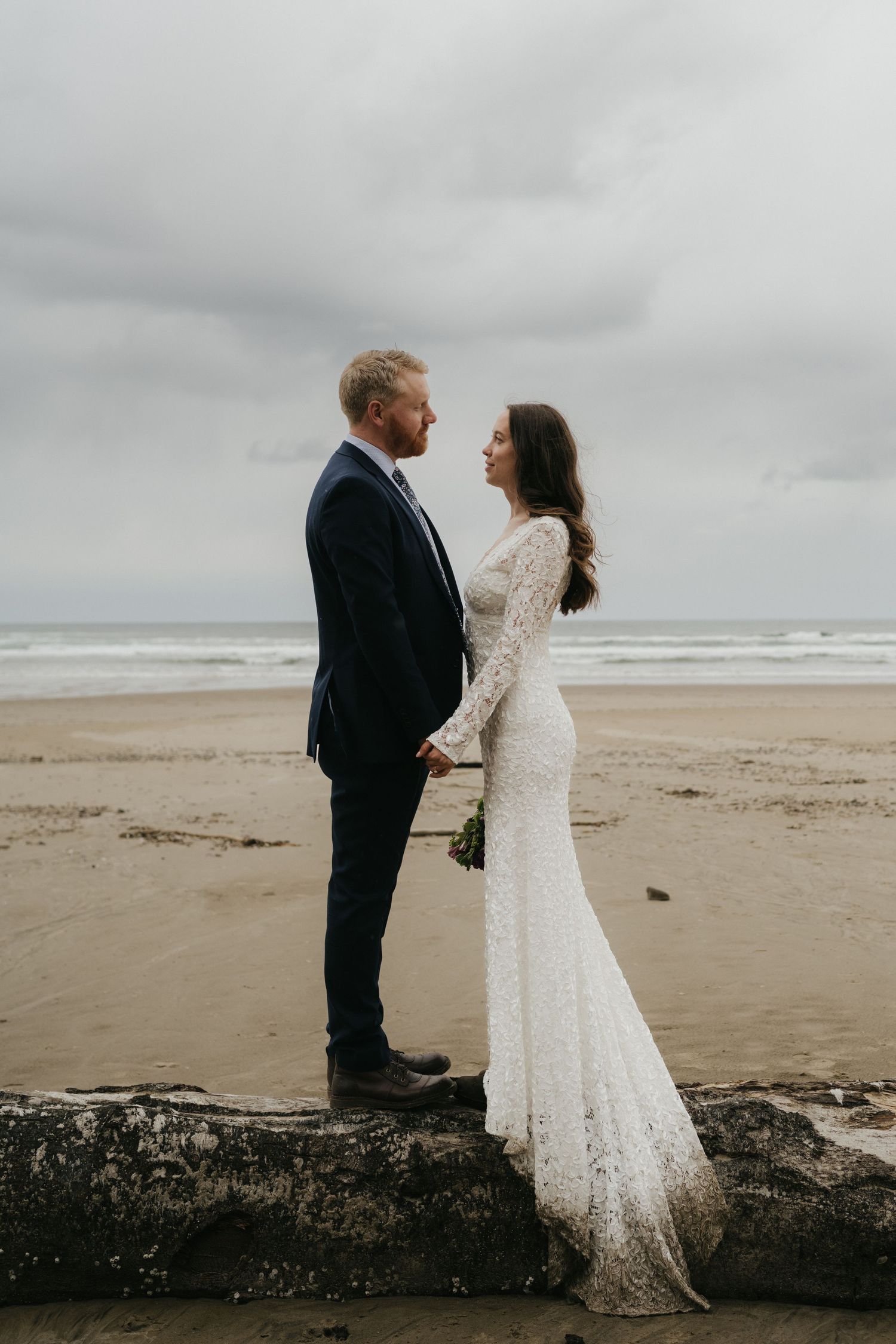 A romantic wedding couple standing on a log at the beach with stormy skies behind them.