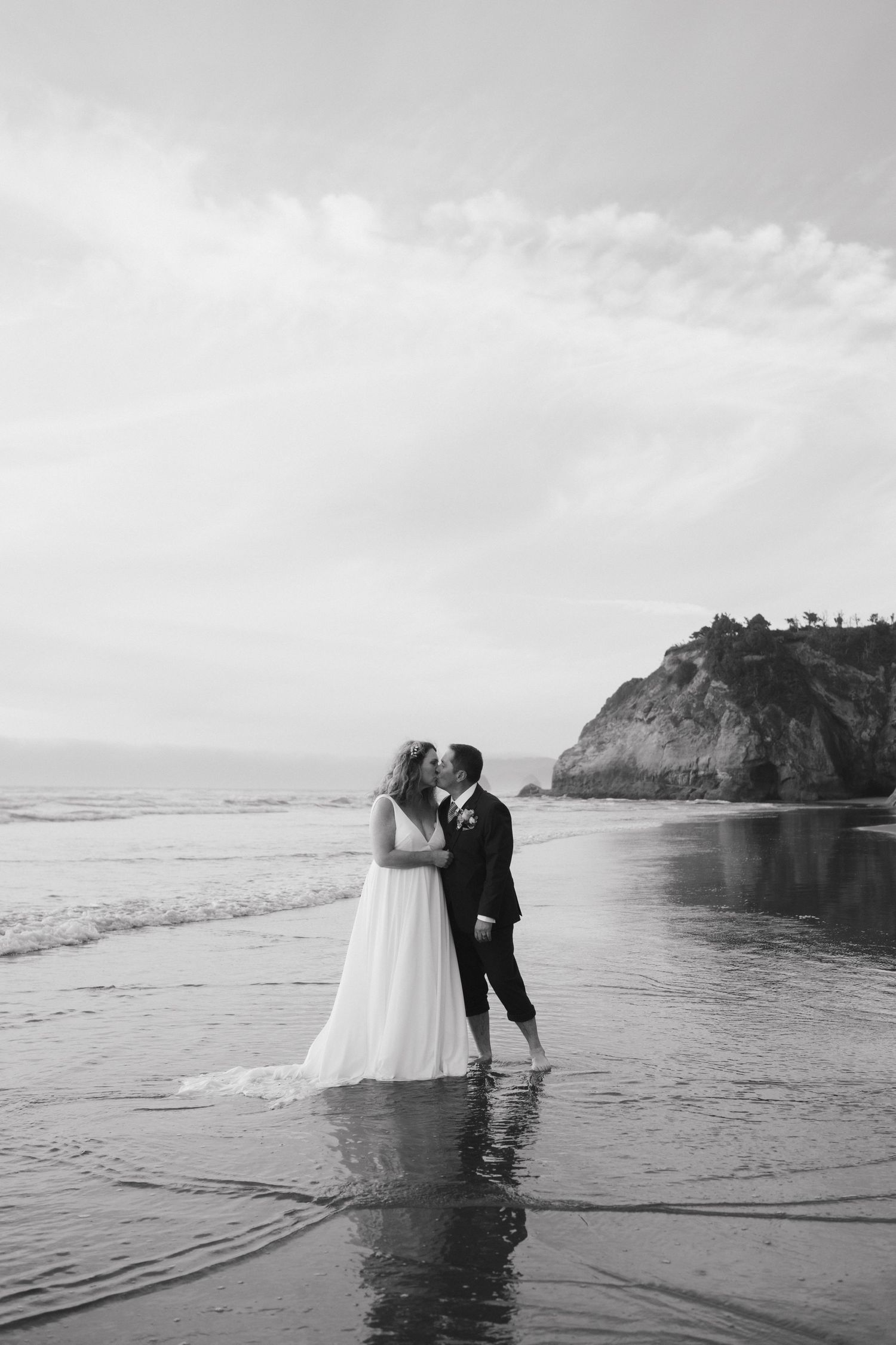 Black and white wedding photo of couple standing in ocean waves with cliffs in background.