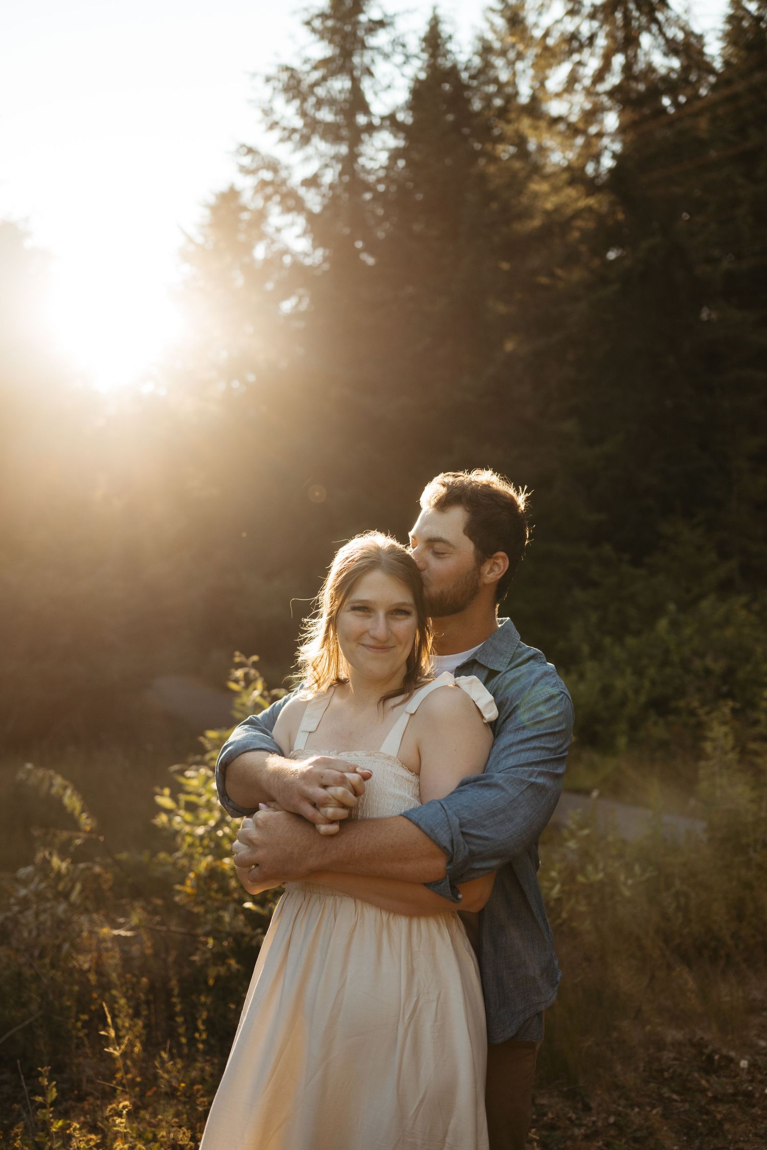 Romantic sunset embrace with couple in cream dress and denim against forest backdrop.