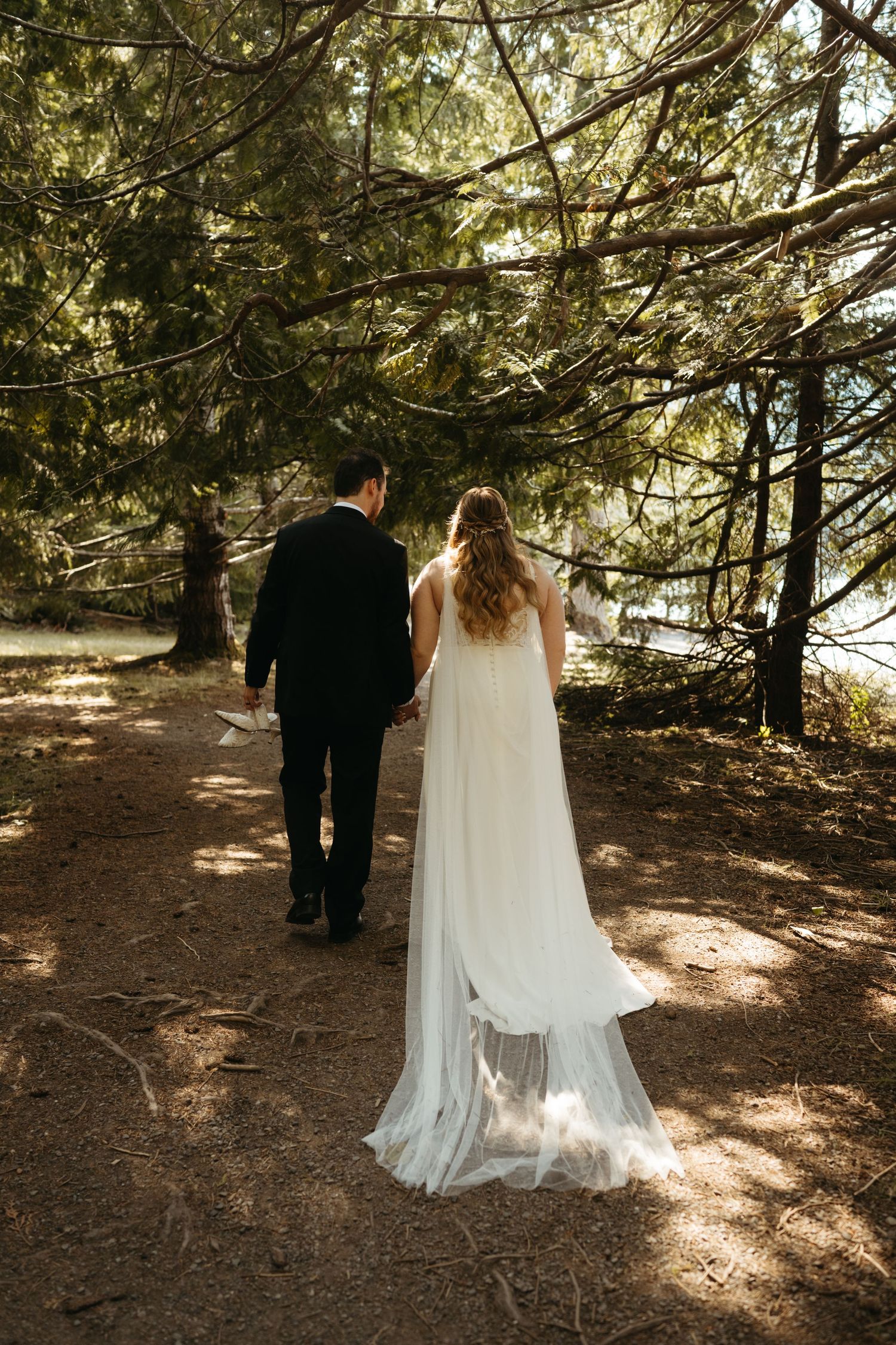 A wedding couple walks hand in hand on a forest path with sunlight filtering through trees.