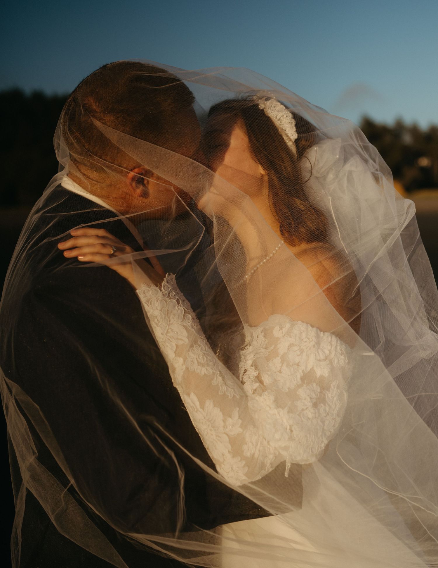 Bride and groom share a romantic kiss under flowing wedding veil in golden hour light.