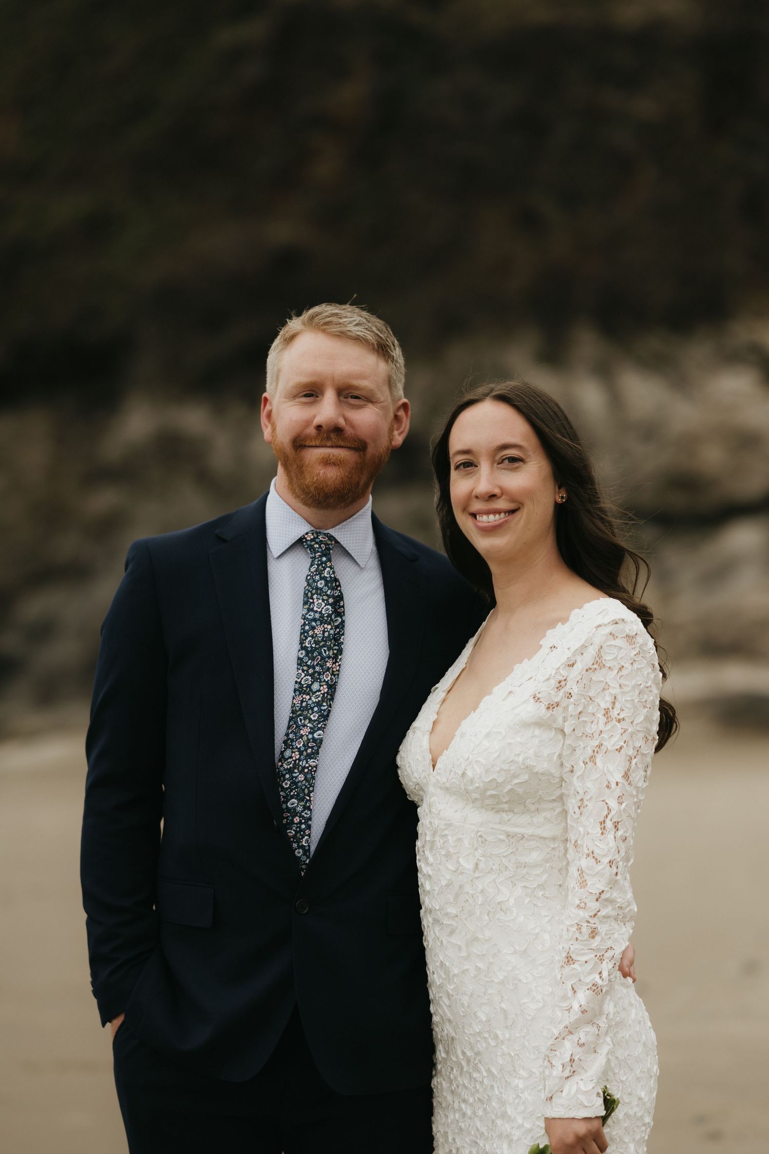 Couple in formal wedding attire smiling on beach with rocky cliffs in background.