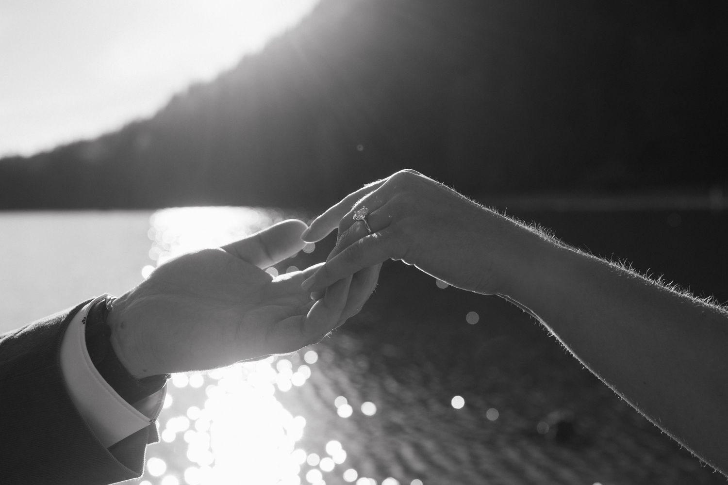 Black and white photo of two hands touching with sunlight and water sparkling in the background.