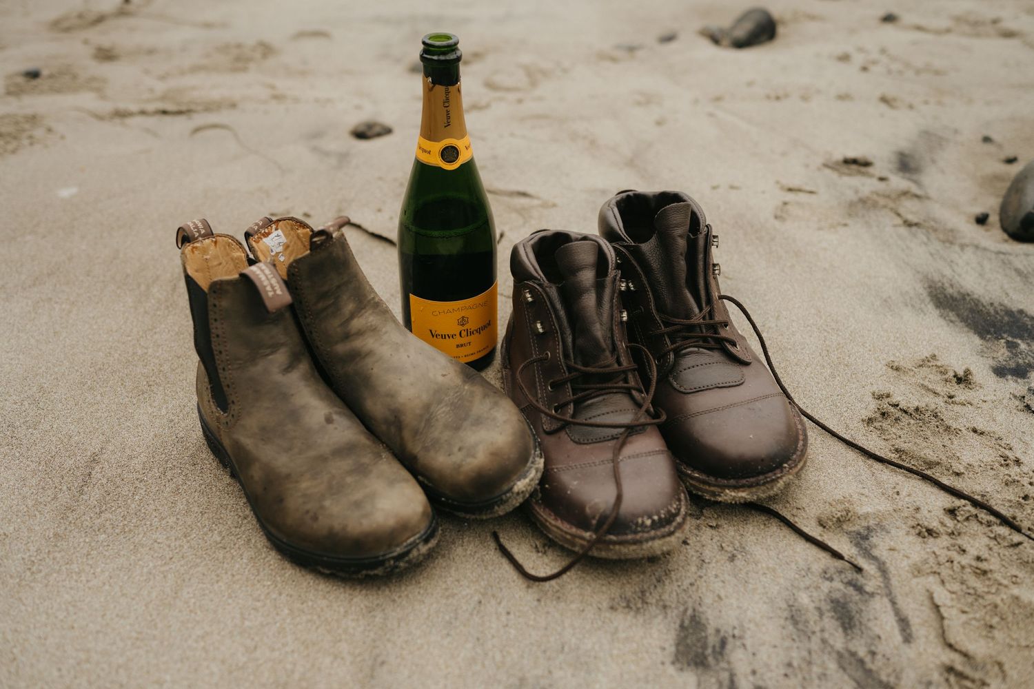 Two pairs of worn boots and a champagne bottle arranged on sandy beach.