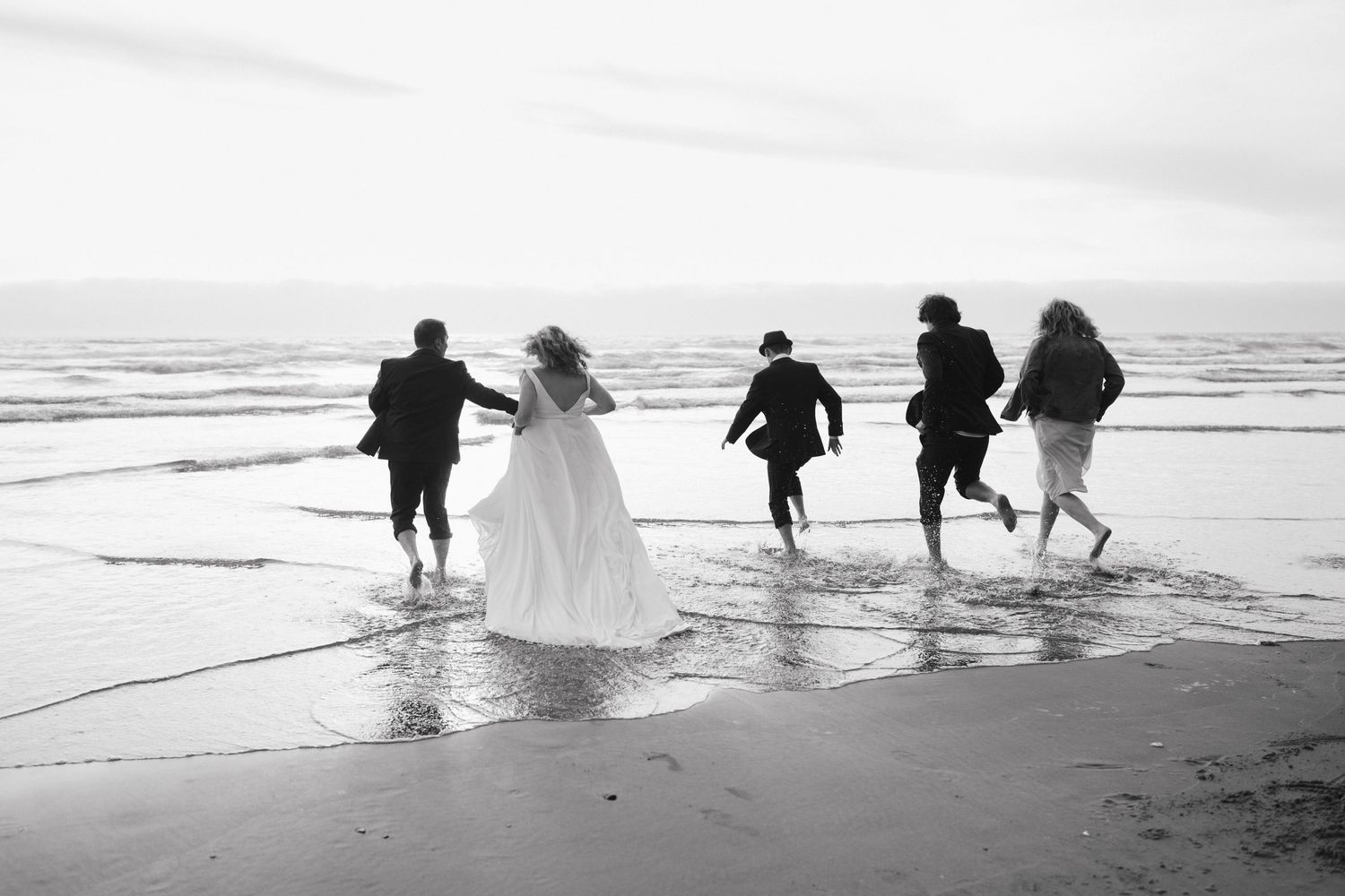 Wedding party runs joyfully through ocean waves in black and white photography.