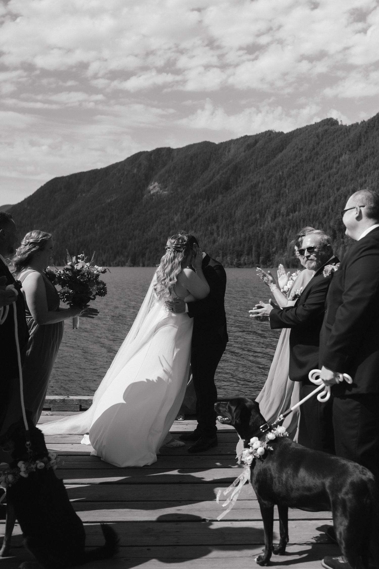 Black and white photo of a lakeside wedding ceremony with a dog in attendance on a wooden dock.