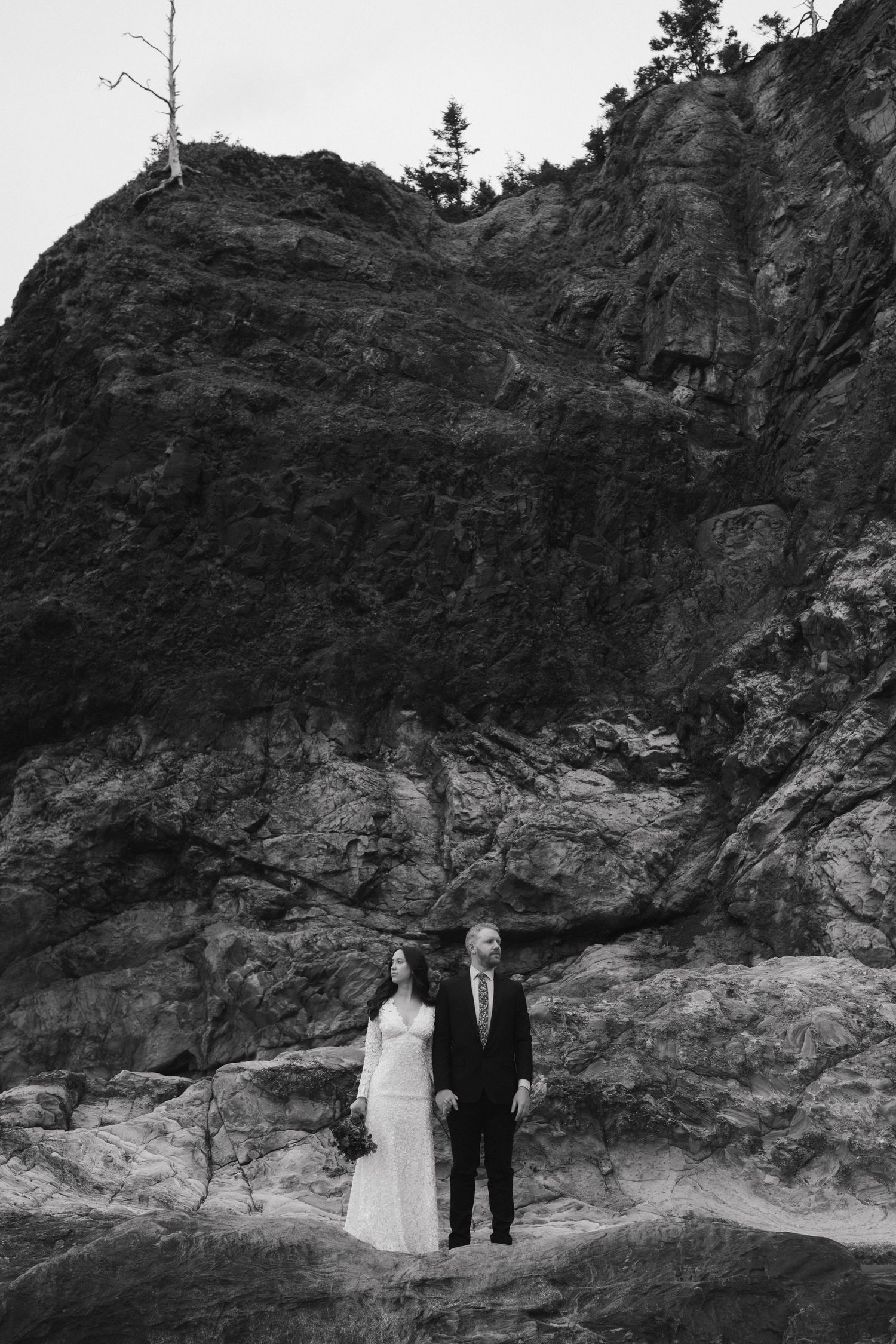 Wedding couple in formal attire standing against dramatic black rock cliff face.