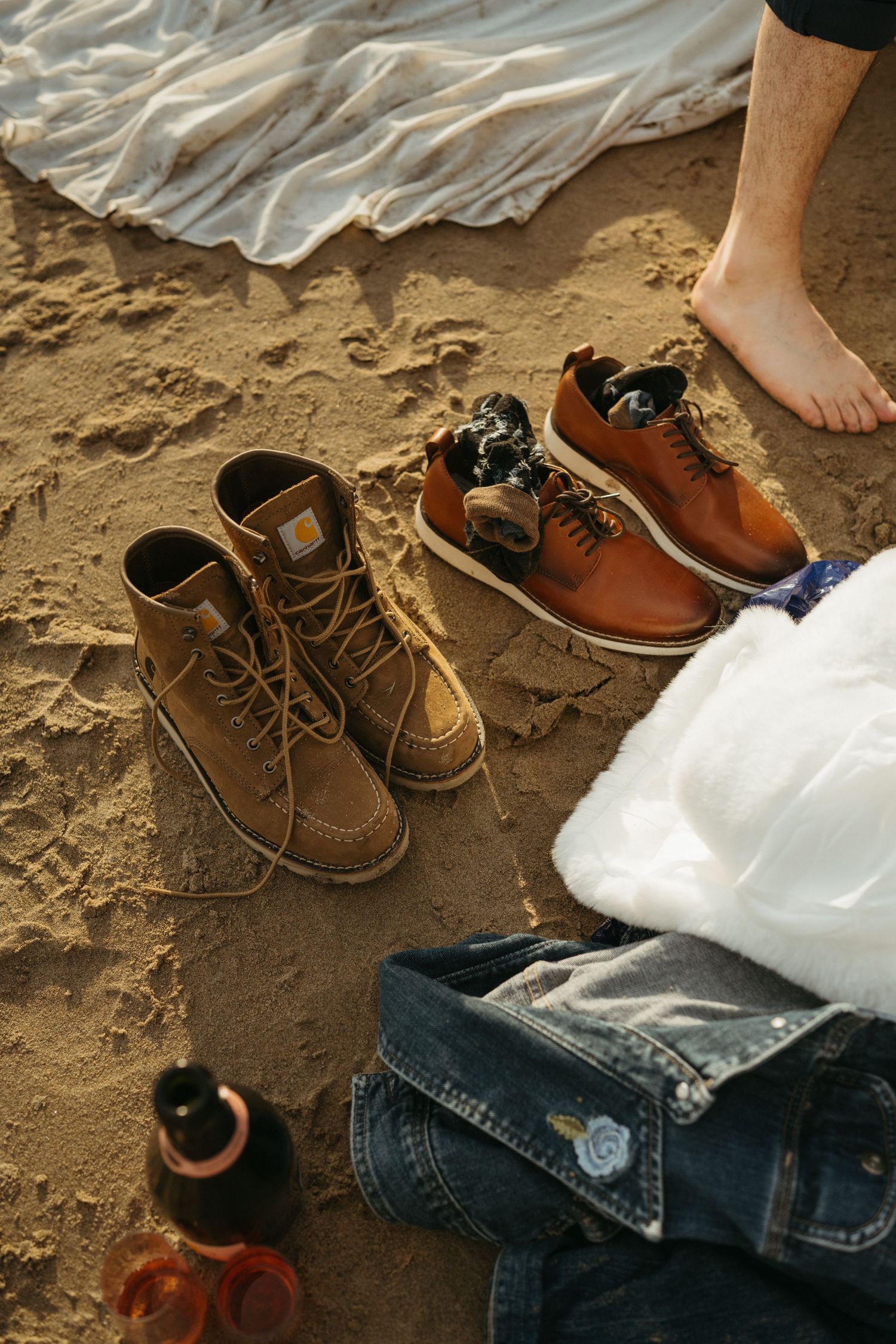 Brown leather boots and shoes arranged on sandy beach next to white dress fabric.