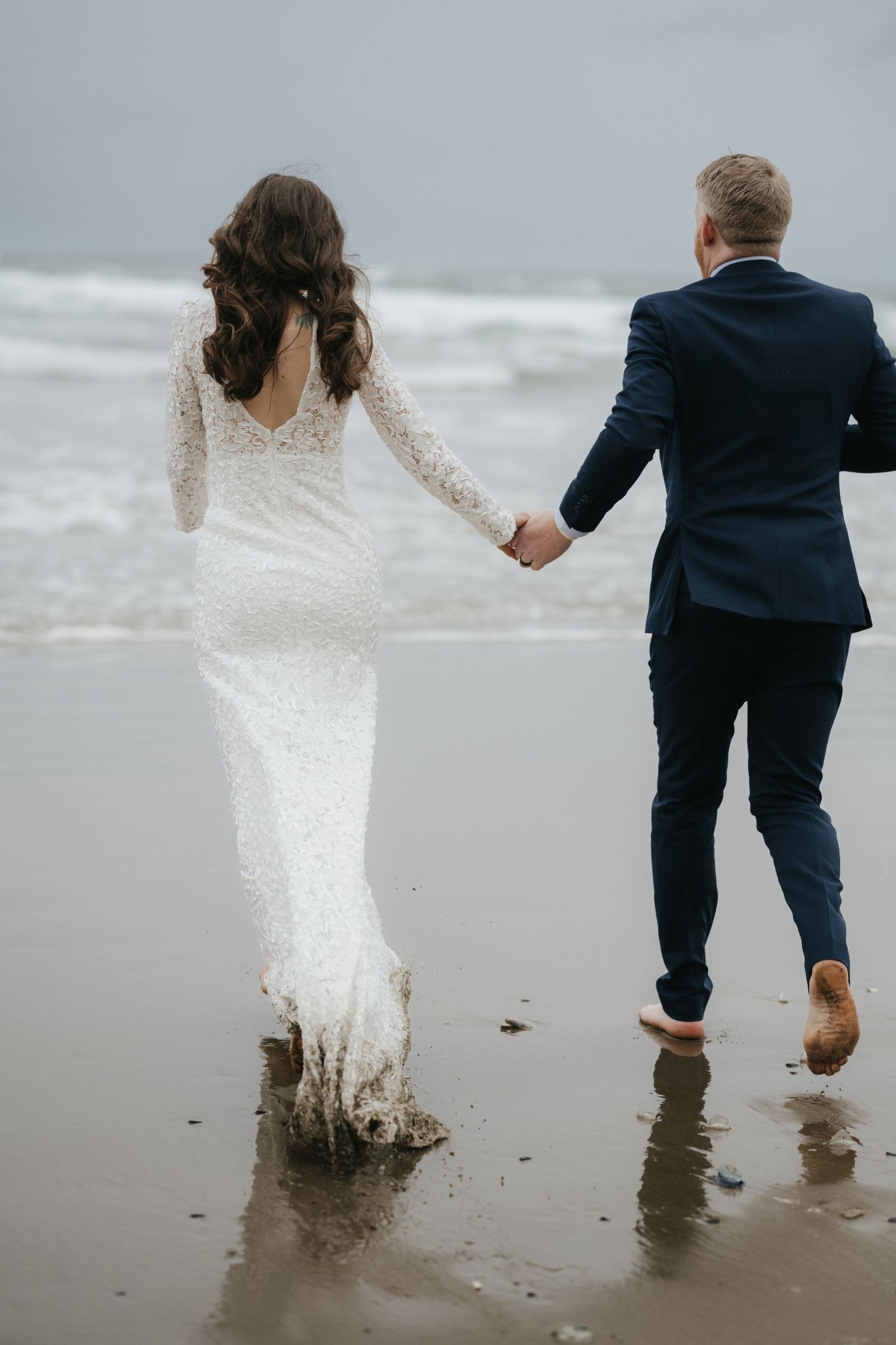Newlyweds walking hand in hand along ocean shore with waves crashing behind them.