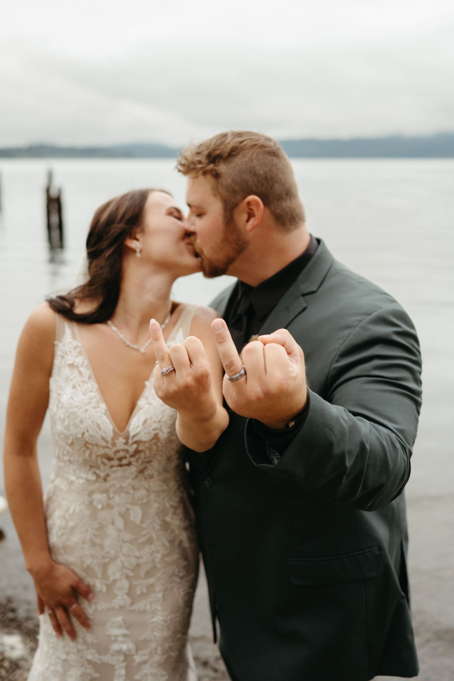 Newlyweds share intimate kiss by waterfront while showing off their rings.