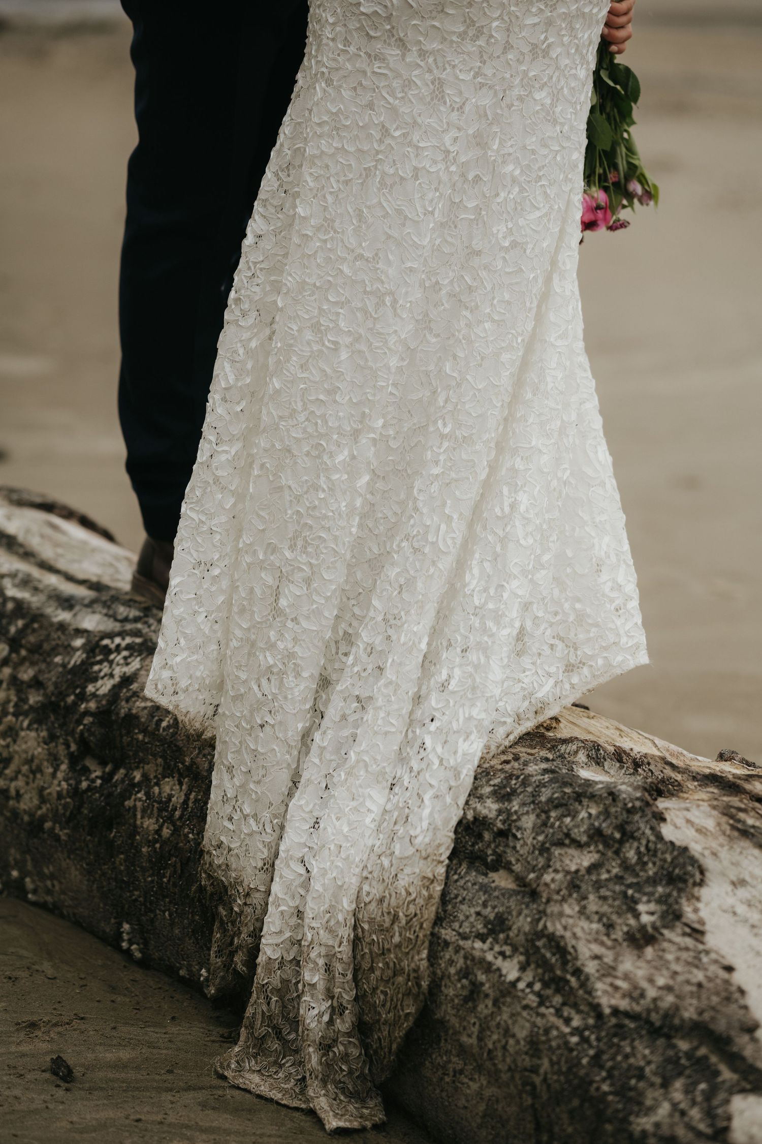 Close-up detail of a lace wedding dress hem against weathered driftwood on a beach.