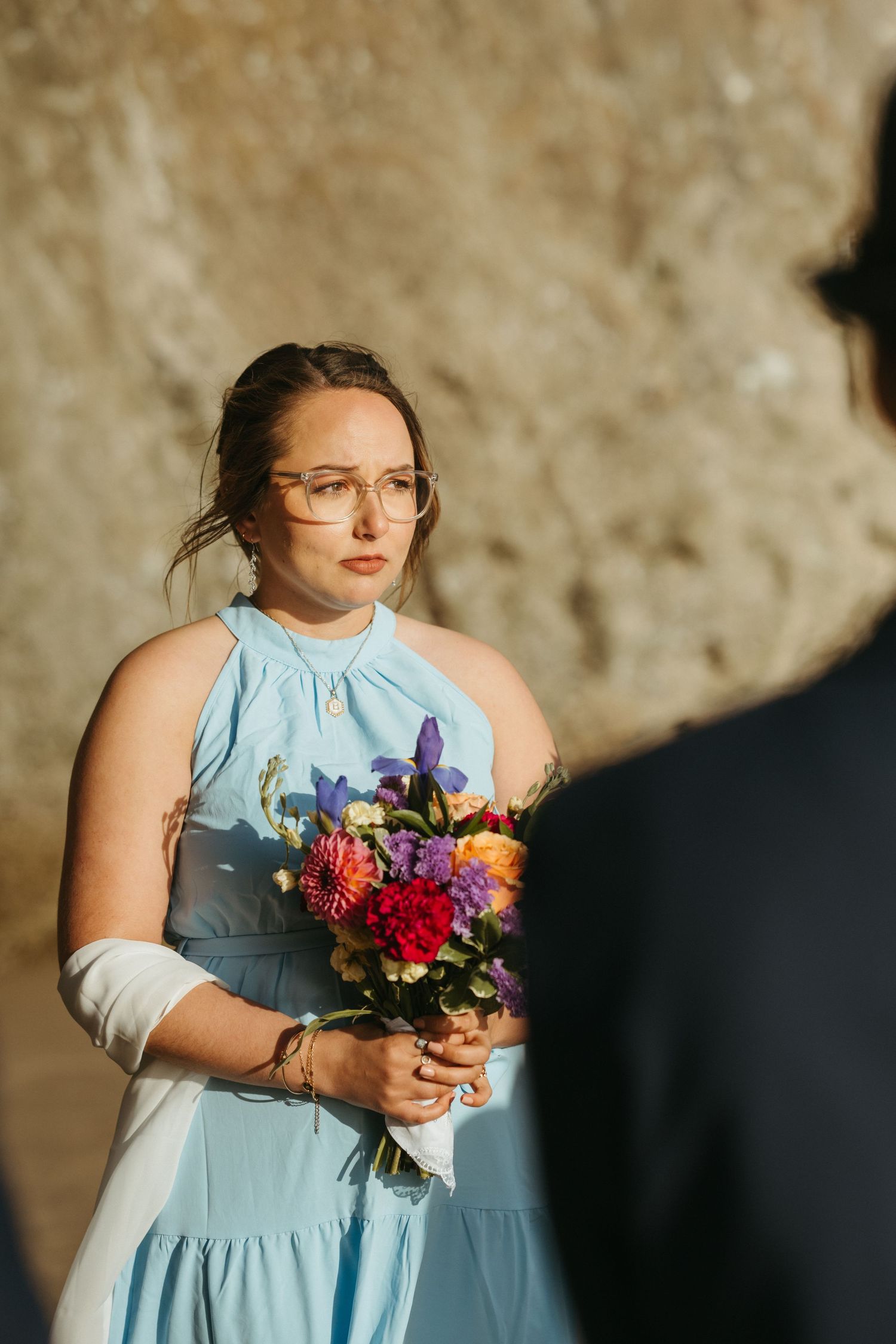 Attendant in light blue dress holds vibrant floral bouquet against stone wall backdrop.