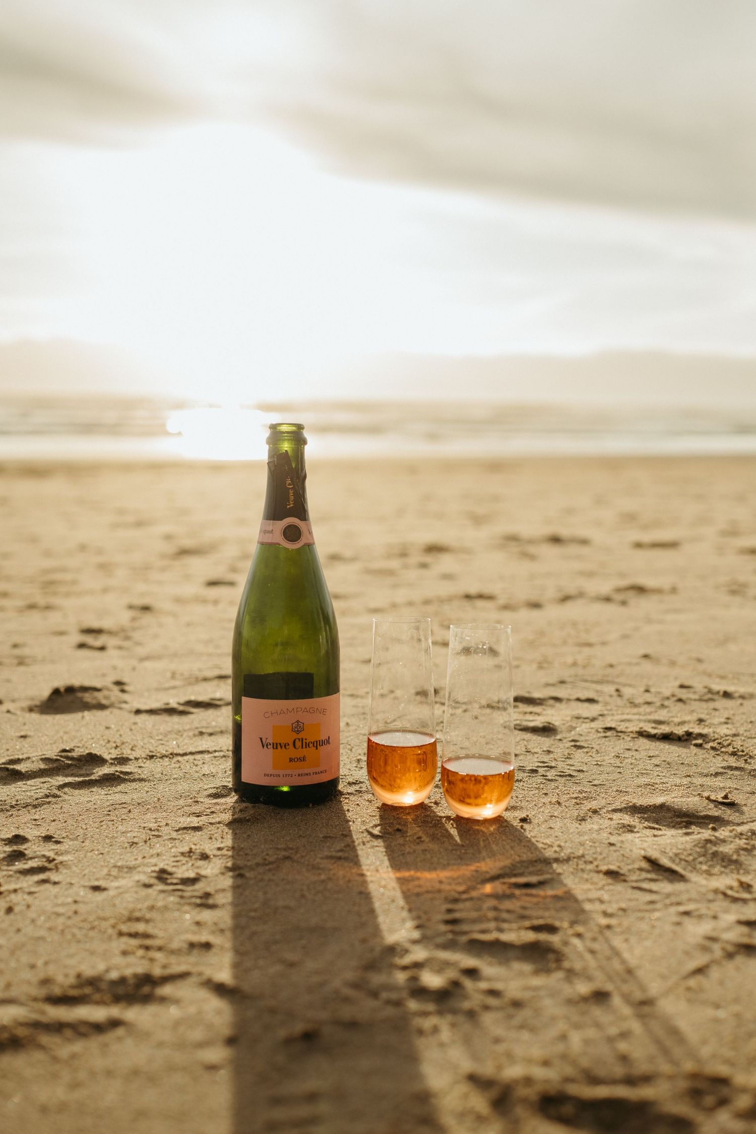 Champagne bottle and glasses set on sandy beach during sunset.