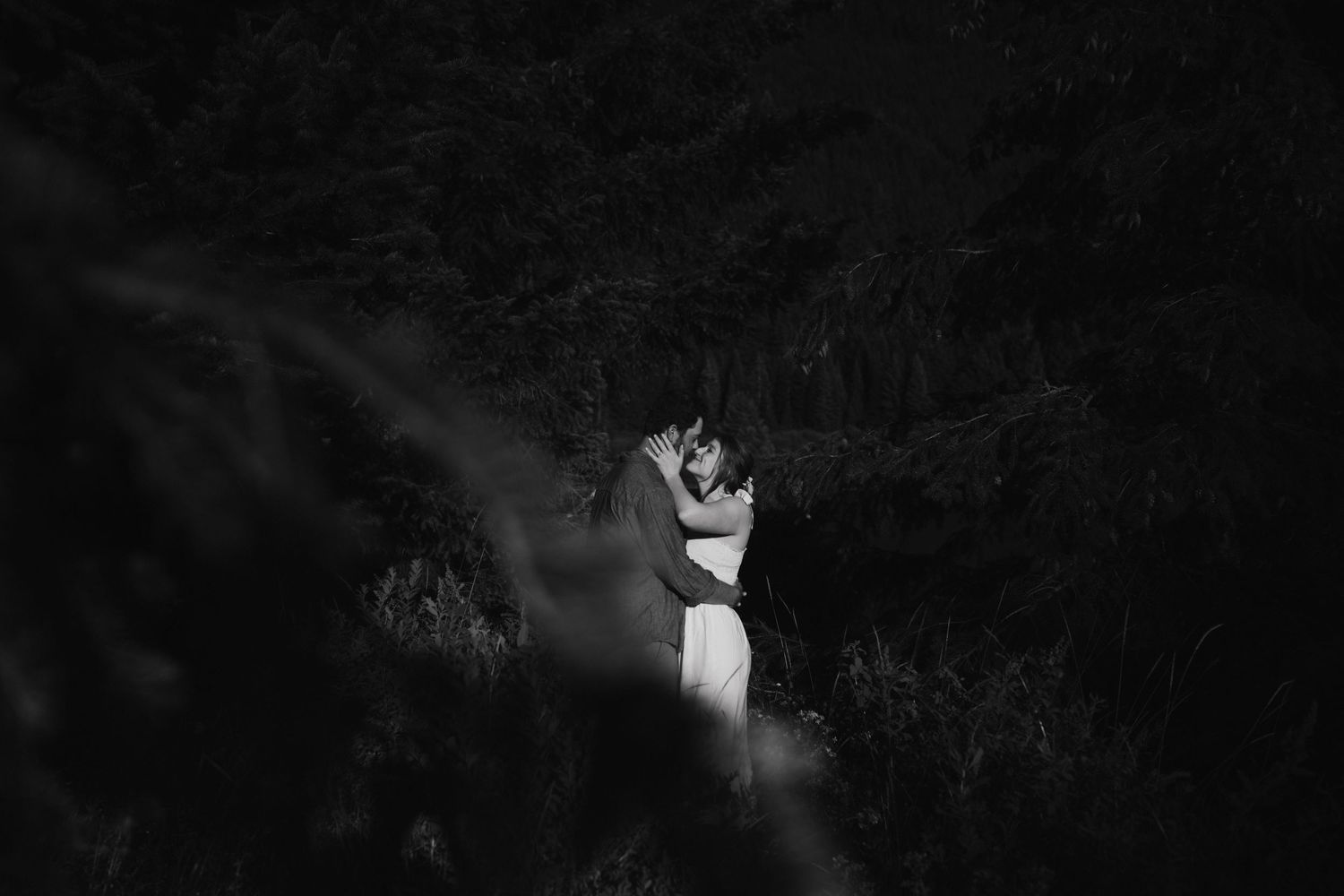 Dramatic black and white photo of couple embracing in moody forest setting.