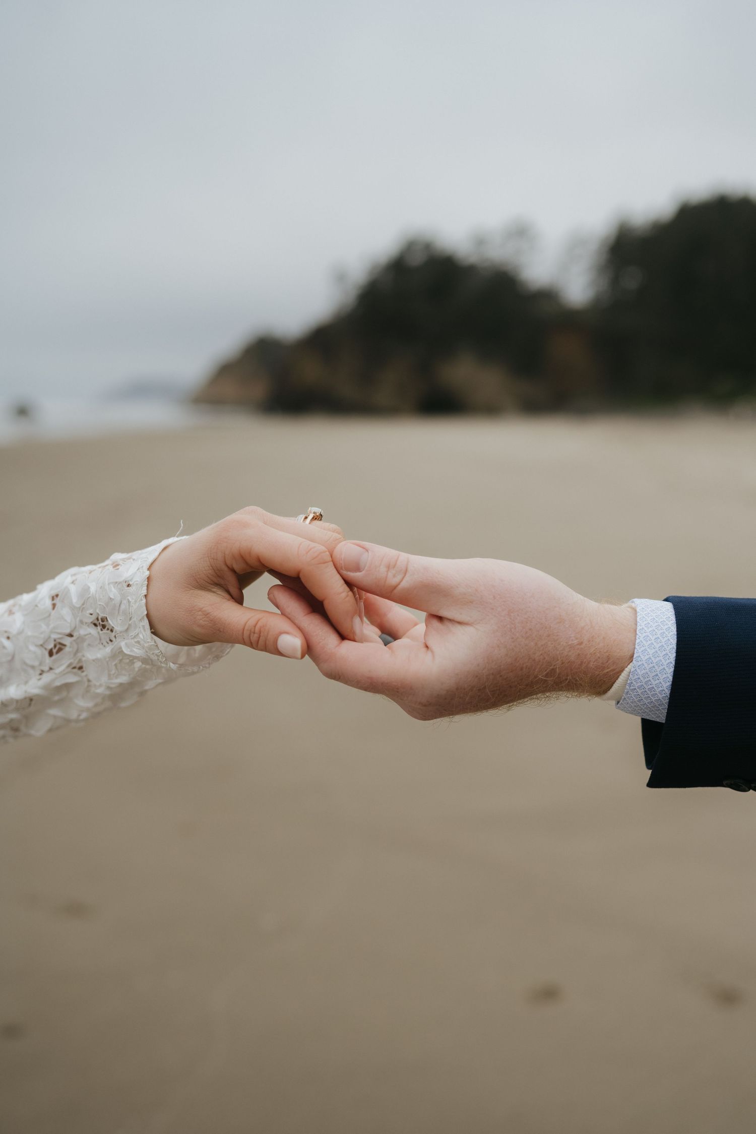Close-up of hands wearing wedding attire reaching toward each other against a beach background.