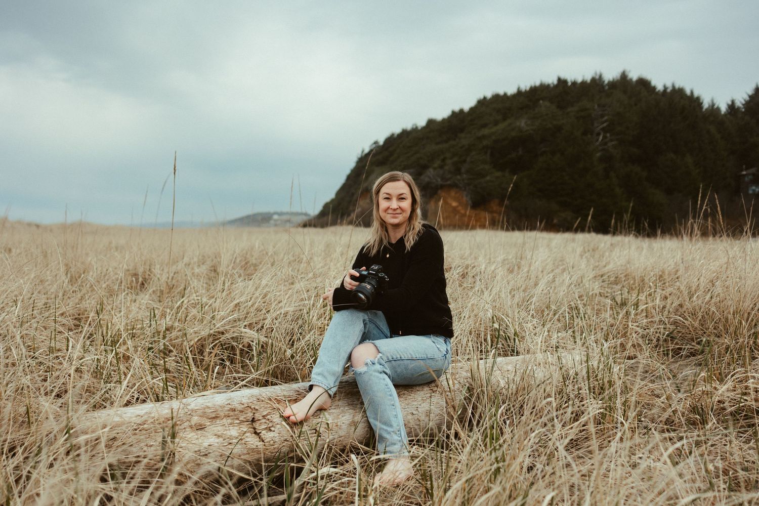 A smiling woman sits on driftwood in a golden grass field near a coastal forest, holding a camera on a cloudy day.
