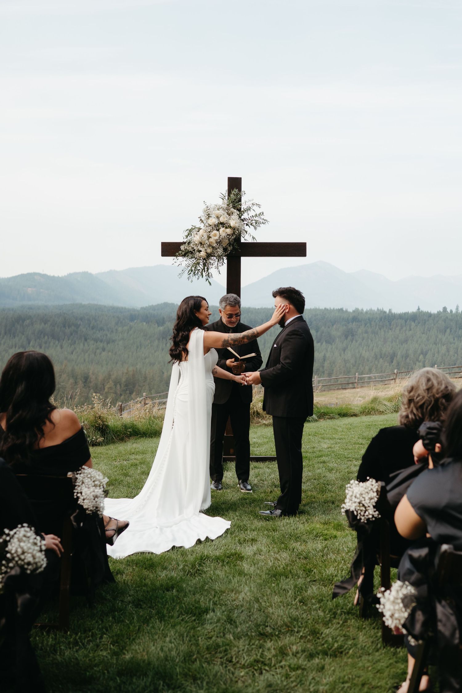 Outdoor mountain wedding ceremony with wooden cross altar and seated guests on grassy hilltop.