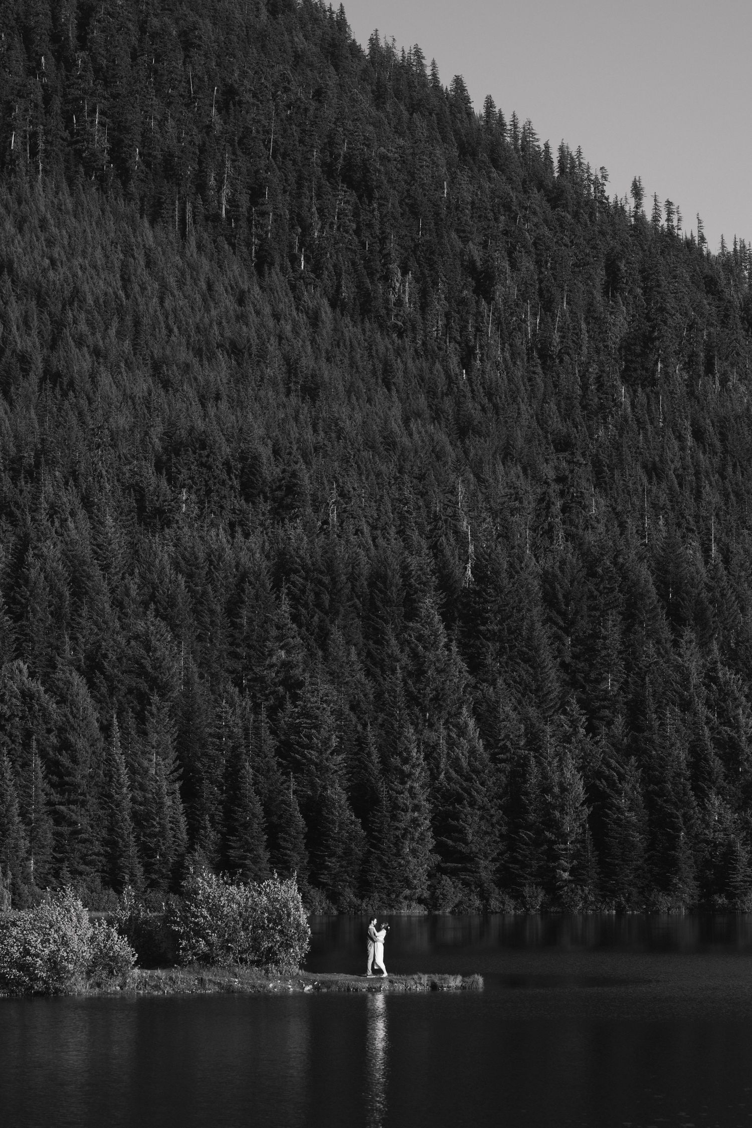 Black and white landscape with couple standing on lake shore against mountainous forest backdrop.
