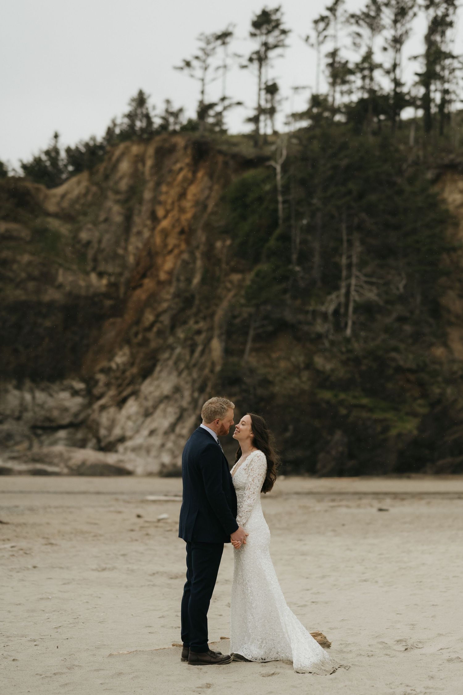 Wedding couple embracing on a beach with dramatic coastal cliffs and pine trees in the background.