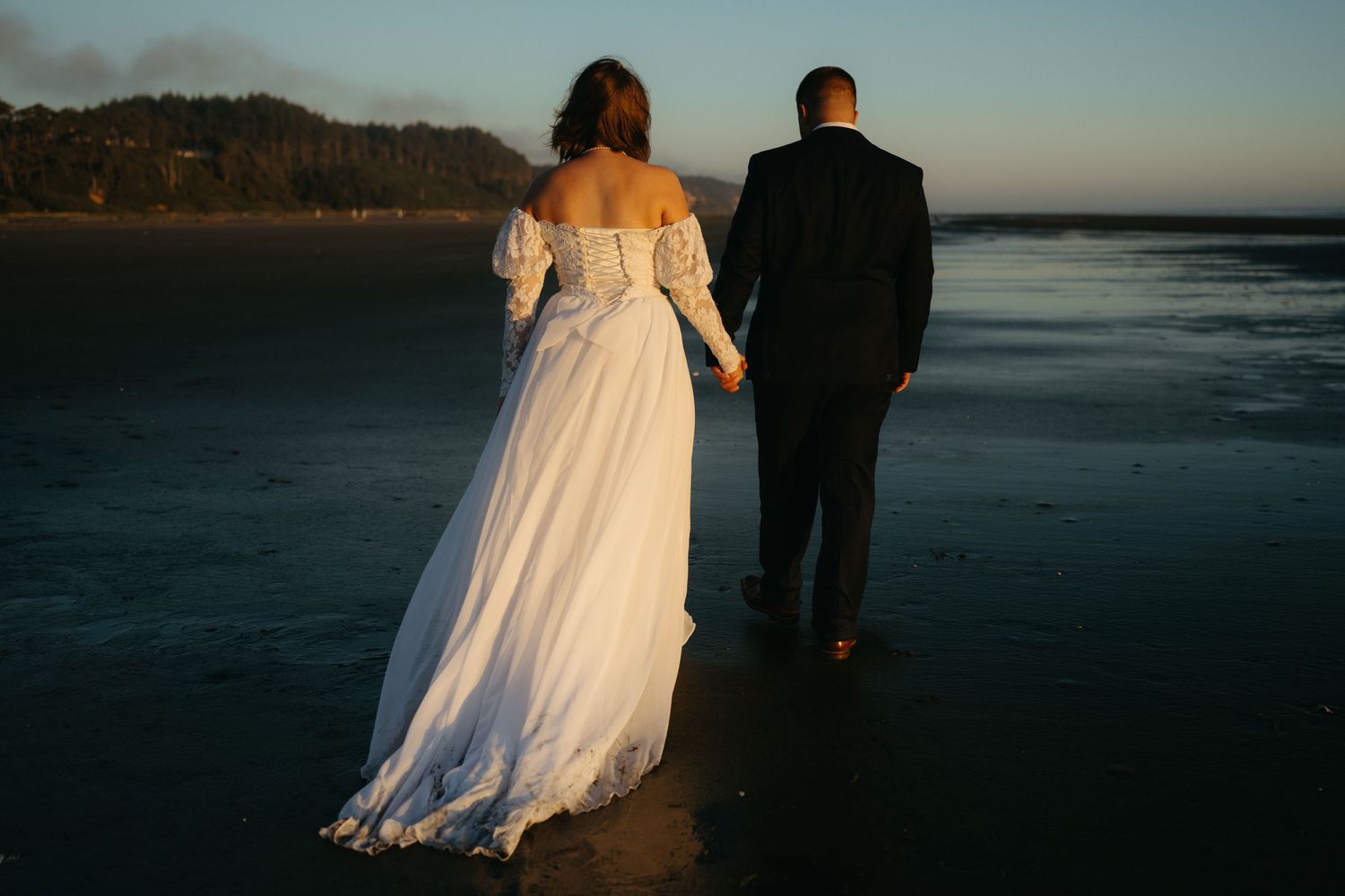 Couple walking along beach shoreline at sunset in formal wedding attire.