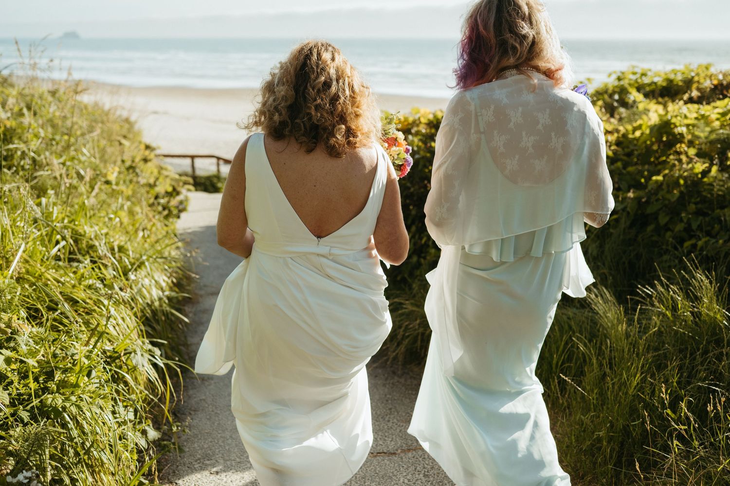 Two people in white dresses walking down coastal path together.