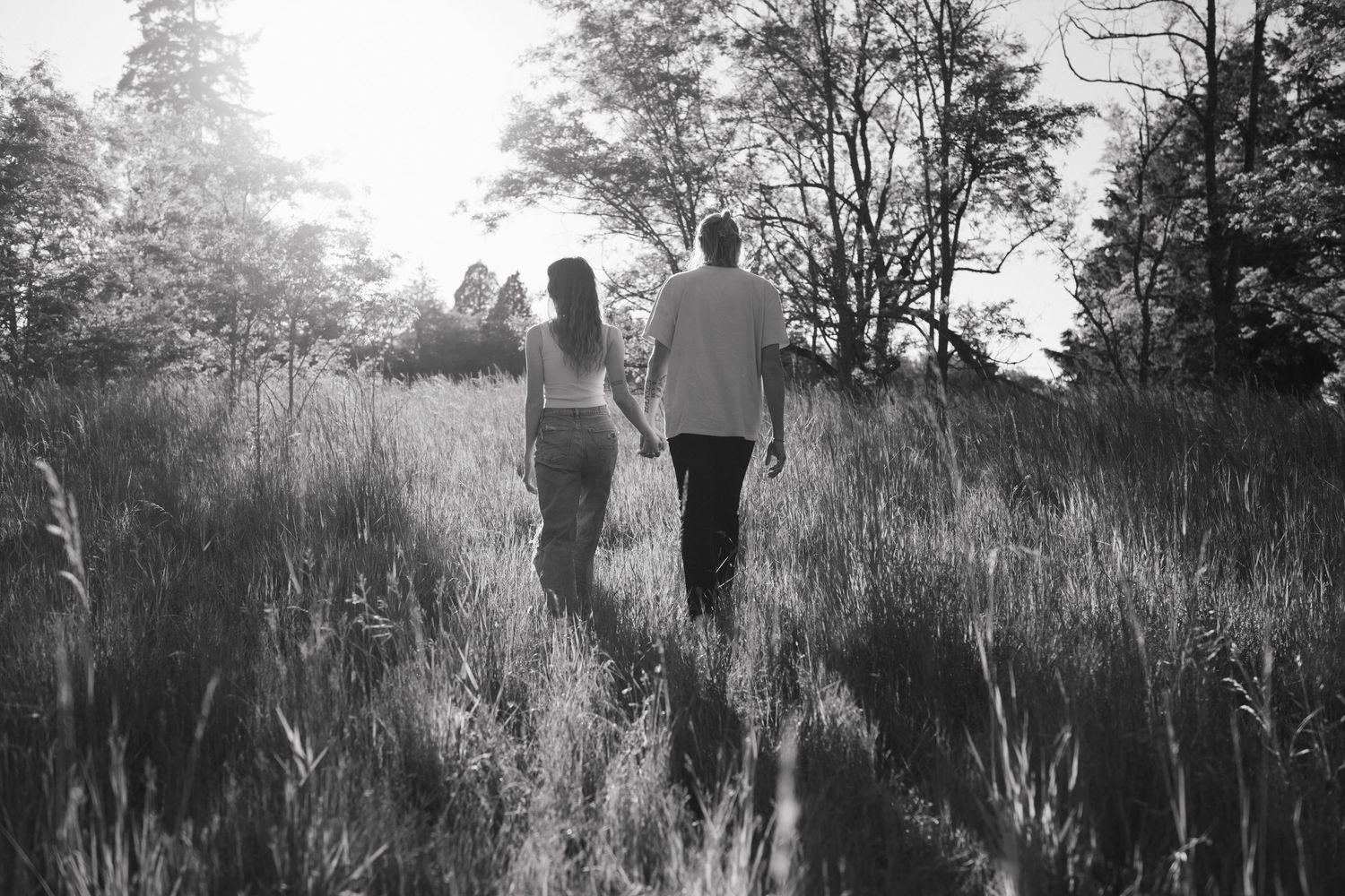Black and white photo of a couple holding hands walking through a sunlit meadow with tall grass and trees.