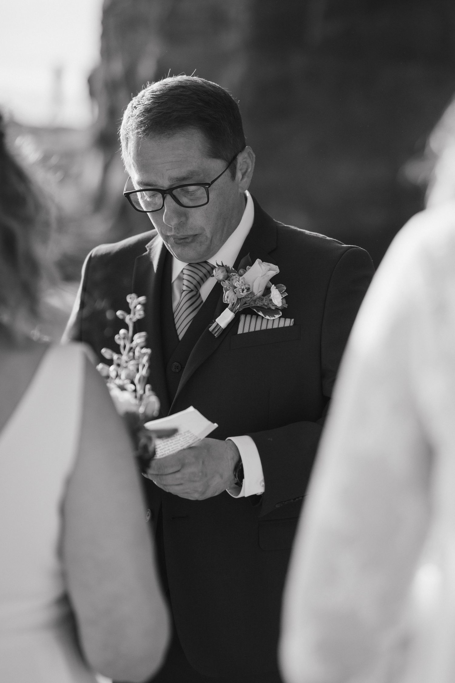 Black and white portrait of groom reading wedding vows while wearing glasses and boutonniere.