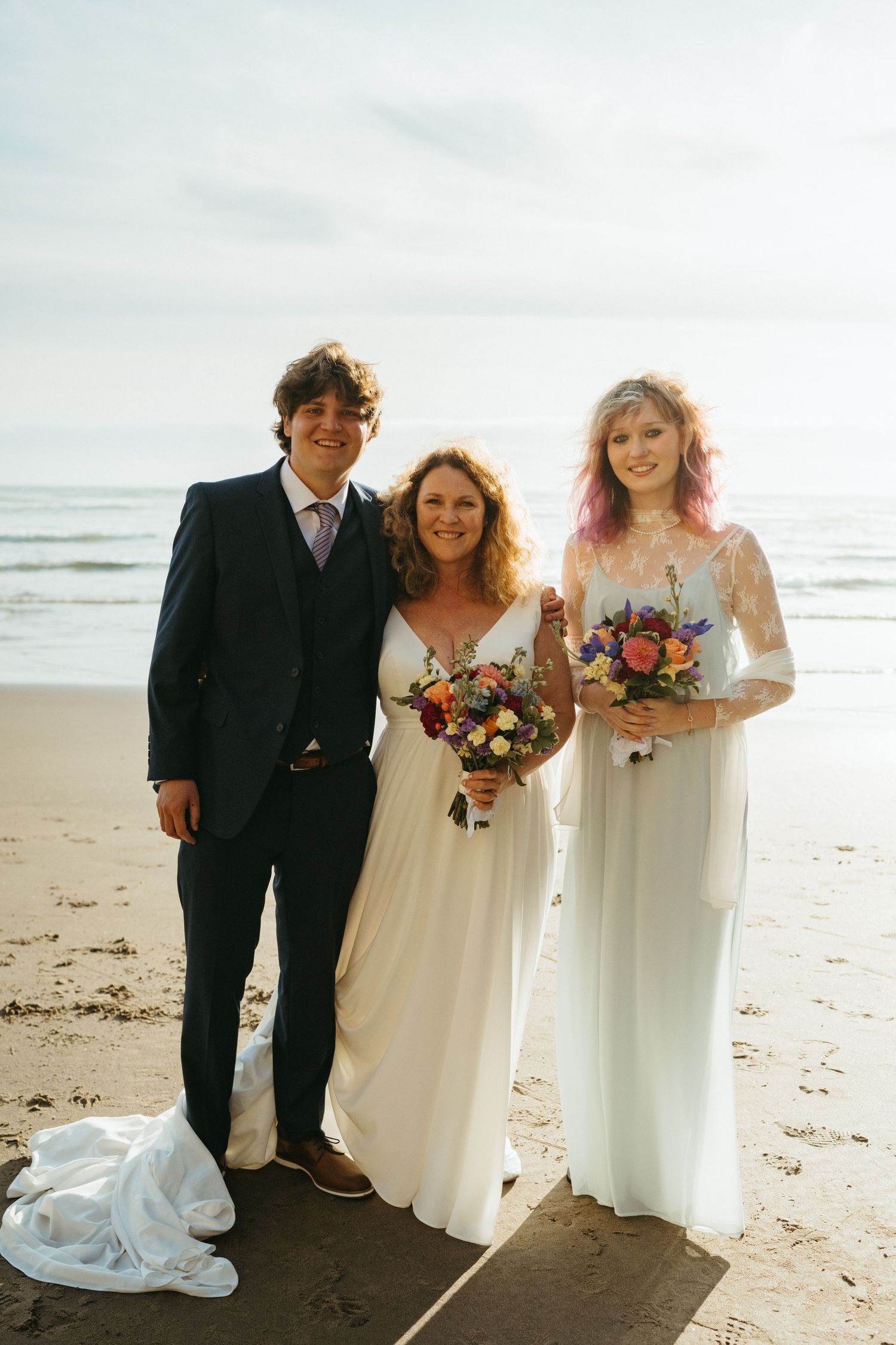 Three wedding attendants pose together with colorful bouquets on the beach.
