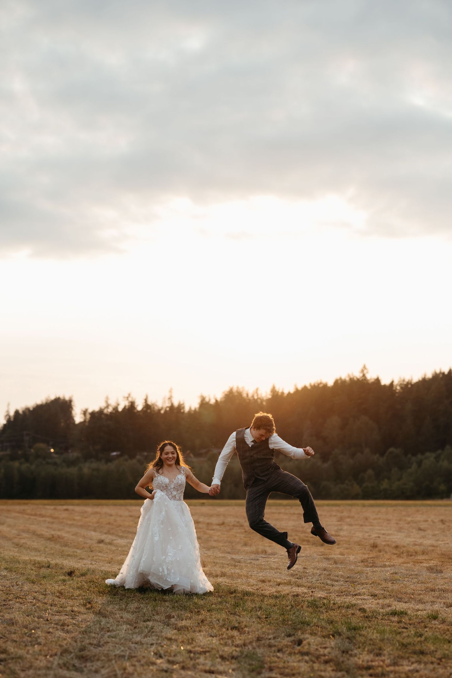 Joyful bride and groom holding hands in a golden field at sunset, groom leaping with excitement in wedding attire.