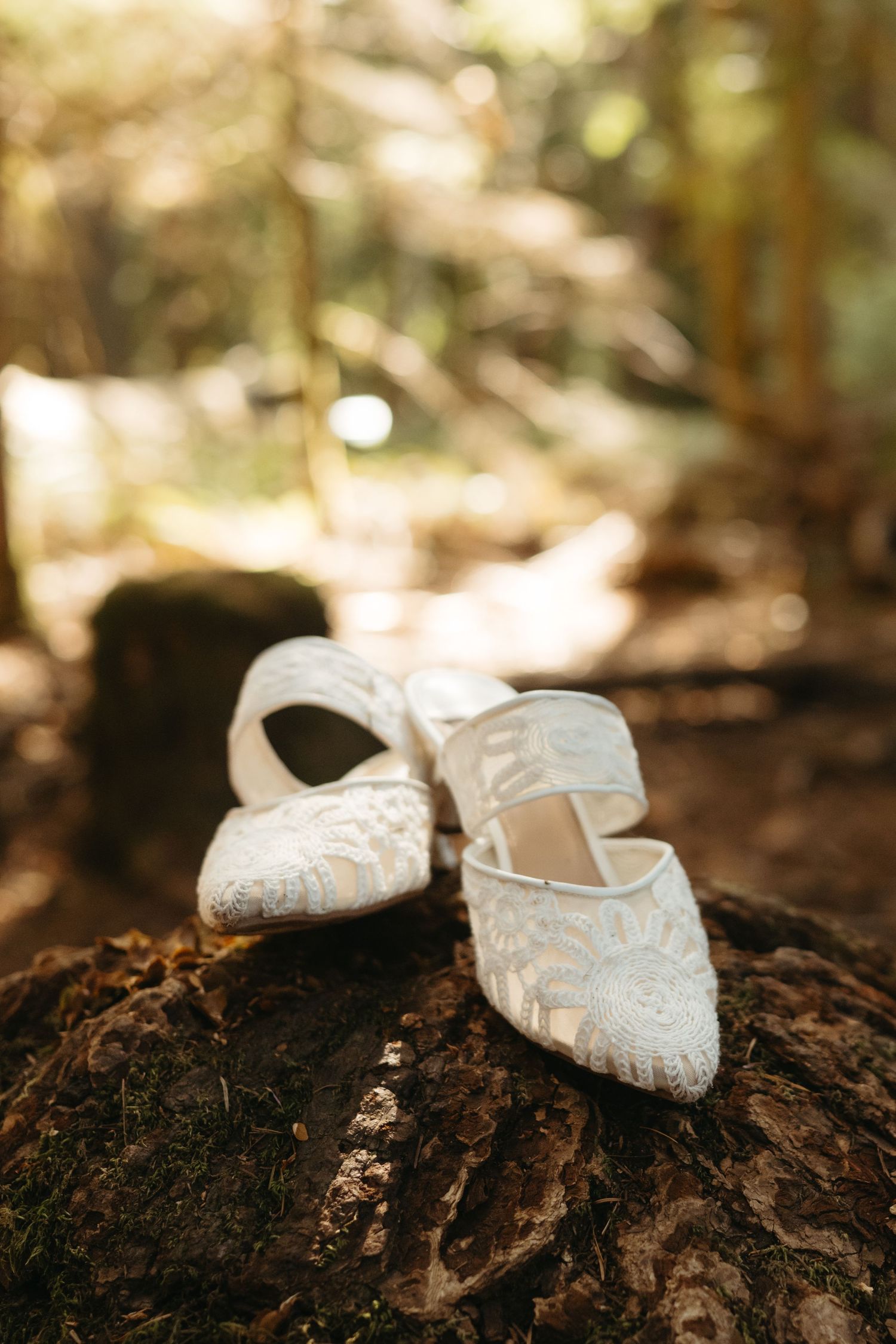 Delicate white lace wedding shoes placed on a rustic wooden log in dappled sunlight.